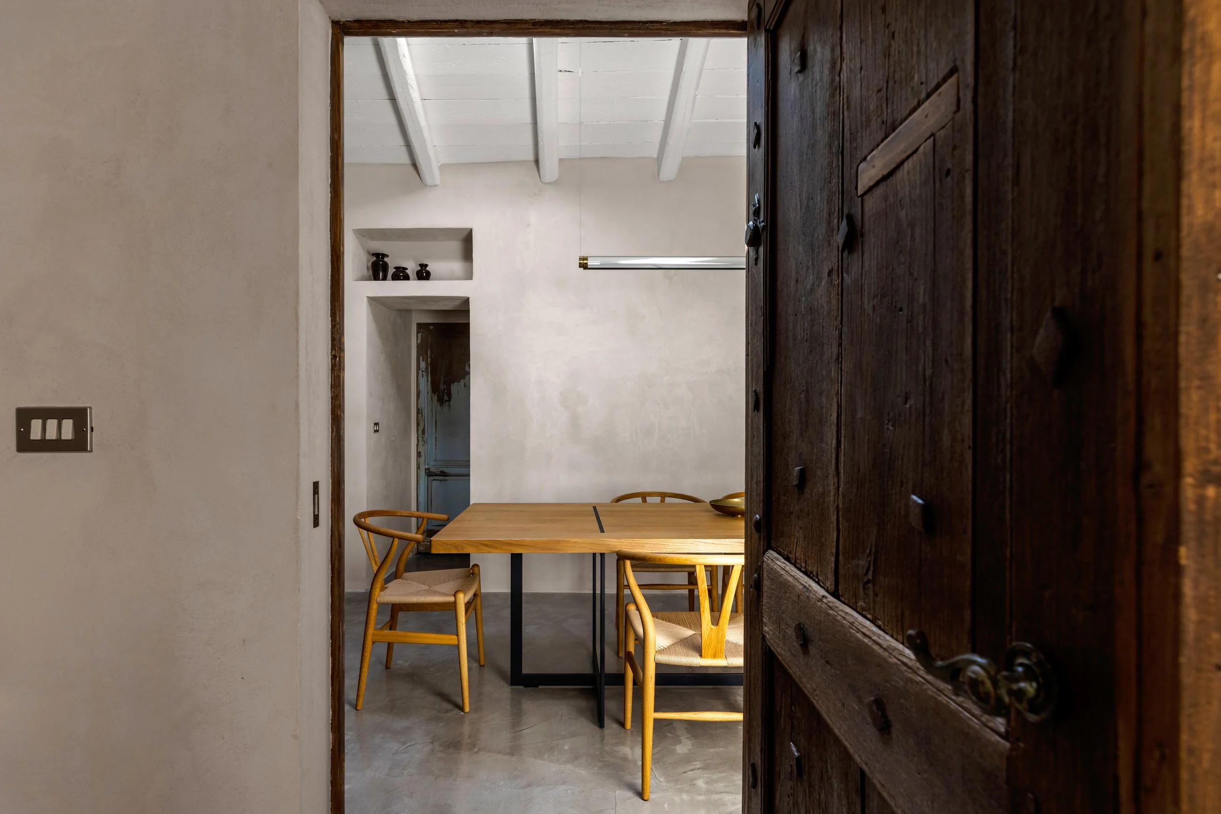 View through a rustic wooden door into a minimalist dining room with a wooden table and four chairs, white walls, a concrete floor, and ceiling beams.