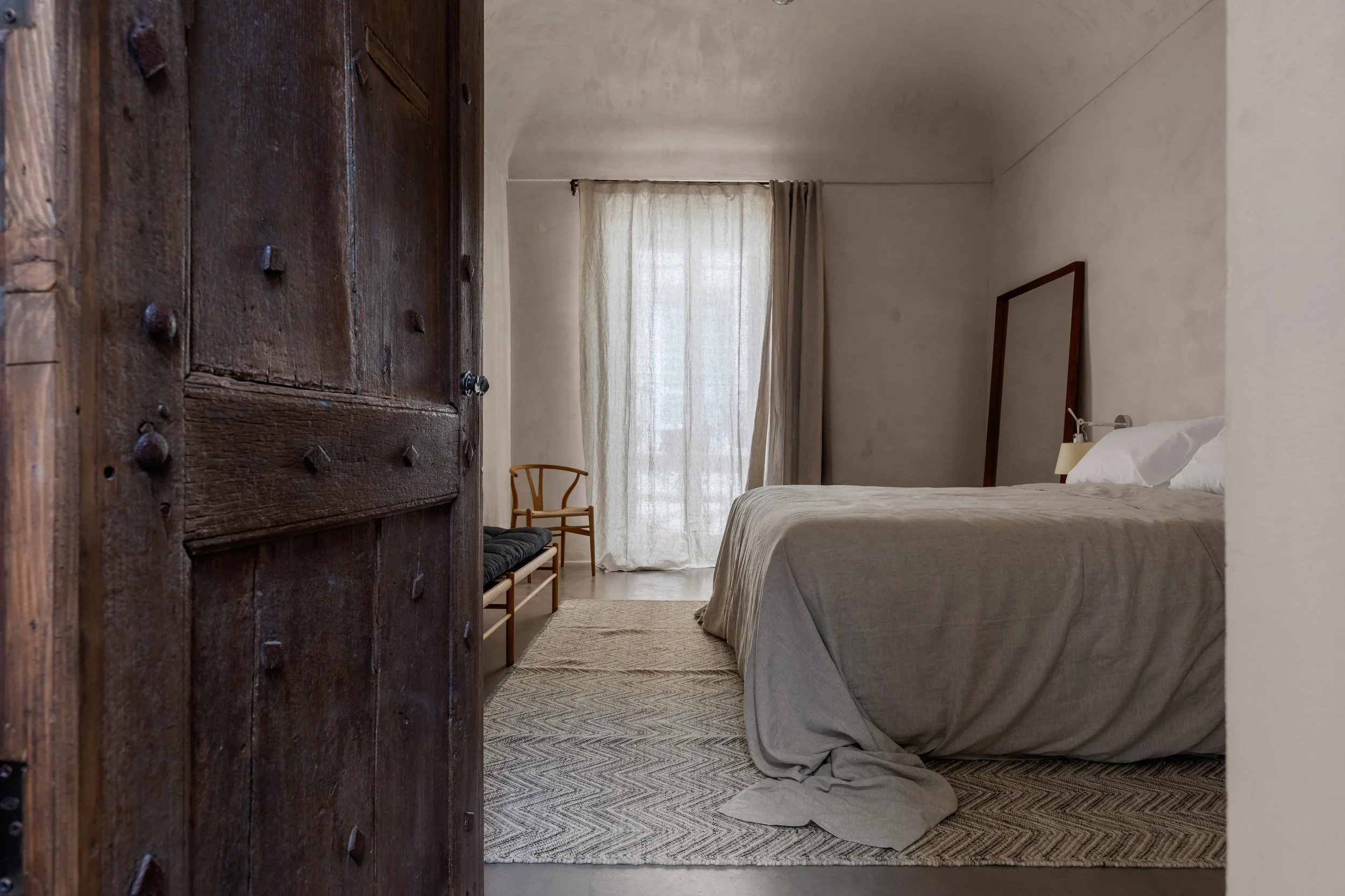 A bedroom with a rustic wooden door, a bed with white sheets, a mirror, a wooden chair, and a window with light curtains.