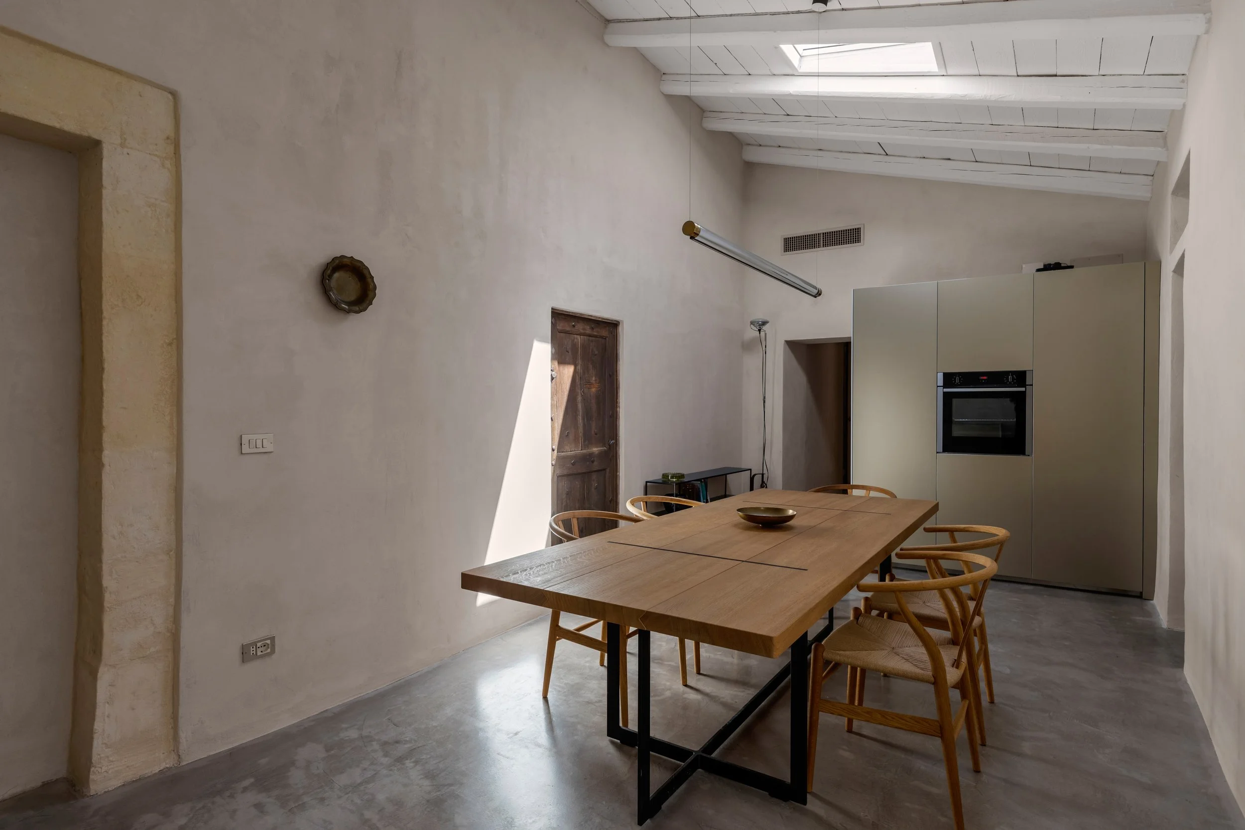 A minimalist dining room with a wooden table and six chairs, a beige wall with a small decorative plate, a wooden door, and a modern kitchen with built-in appliances in the background.