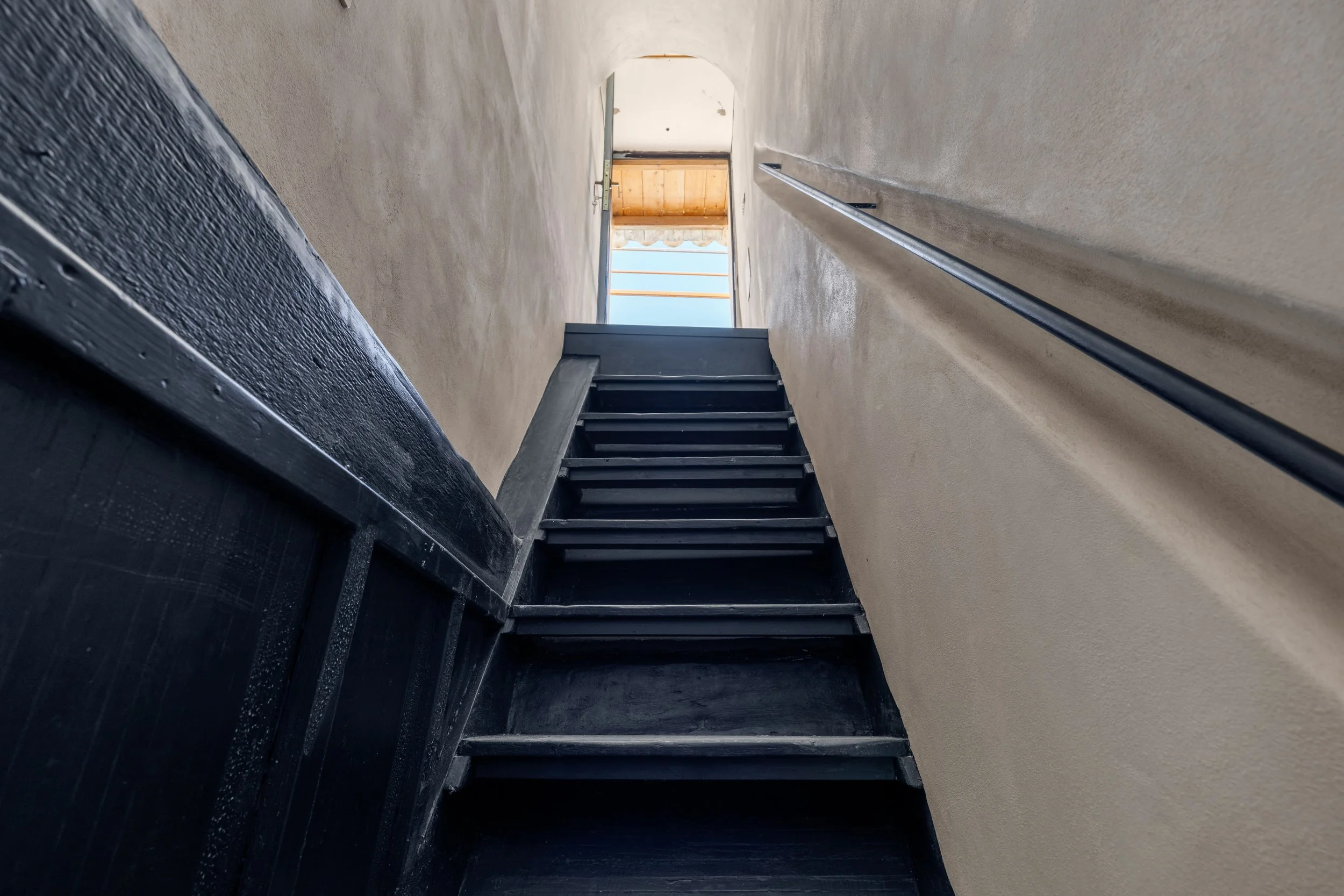 Looking up a narrow staircase with black steps and beige walls, with an open door at the top revealing a balcony or outdoor space.