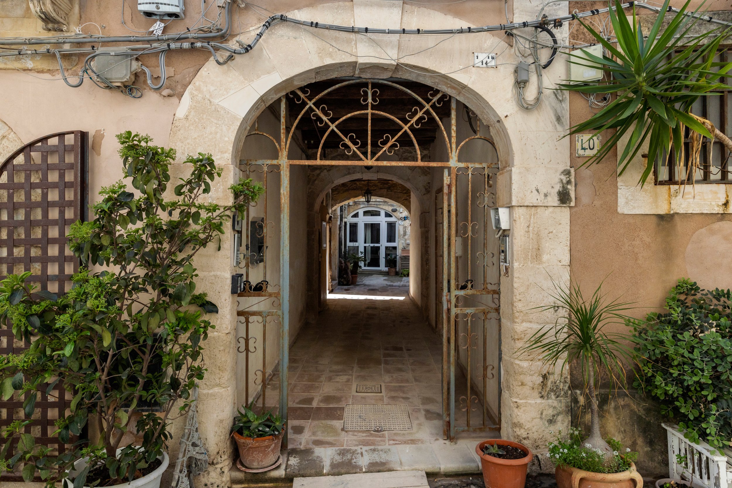 An entrance with a rusty iron gate leading into a courtyard with potted plants and a pathway, surrounded by worn stone walls and electrical wiring.