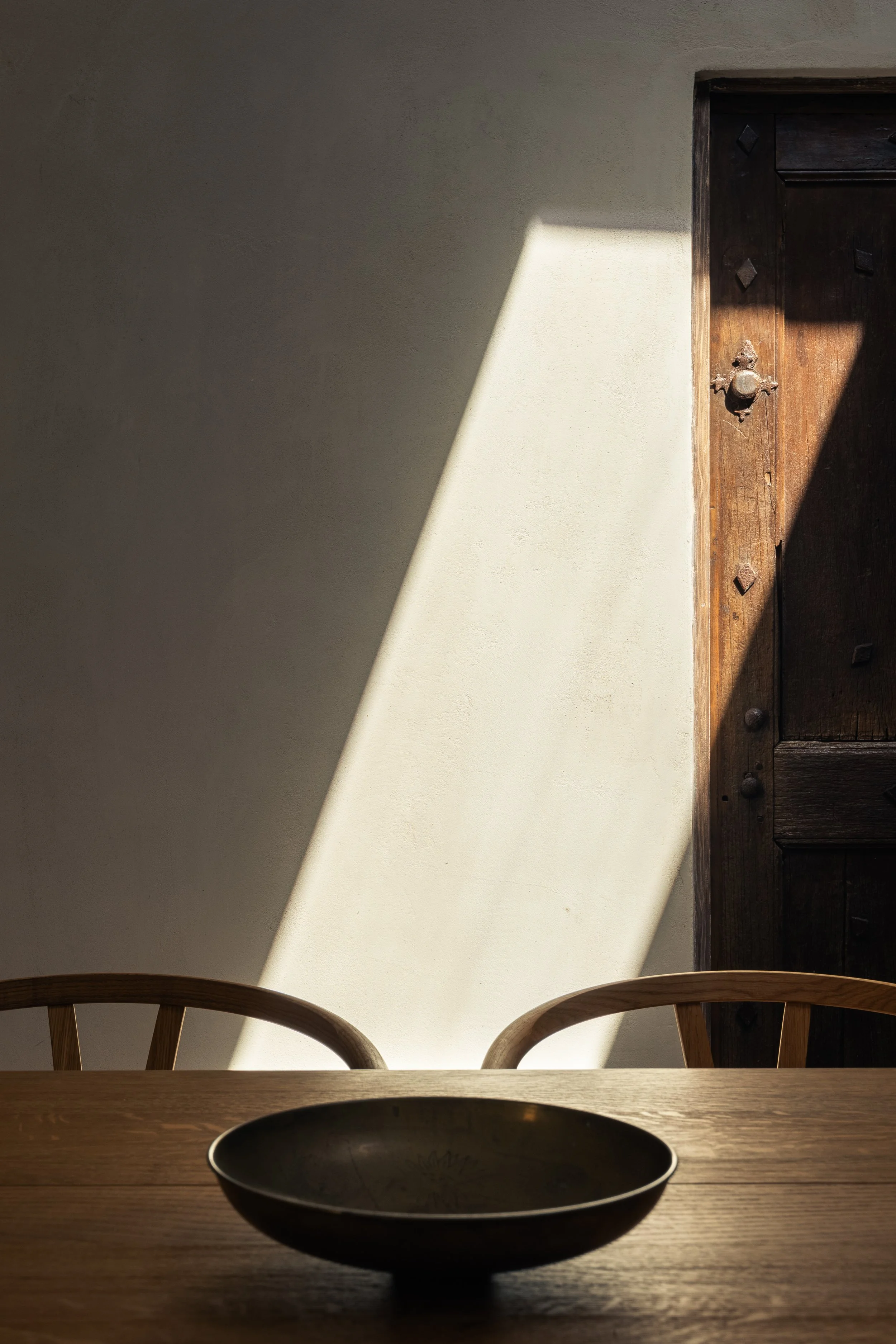 A wooden dining table with a shallow bowl on it, two wooden chairs with curved backs, a textured wall with sunlight forming a shadow, and a dark wooden door in the background.