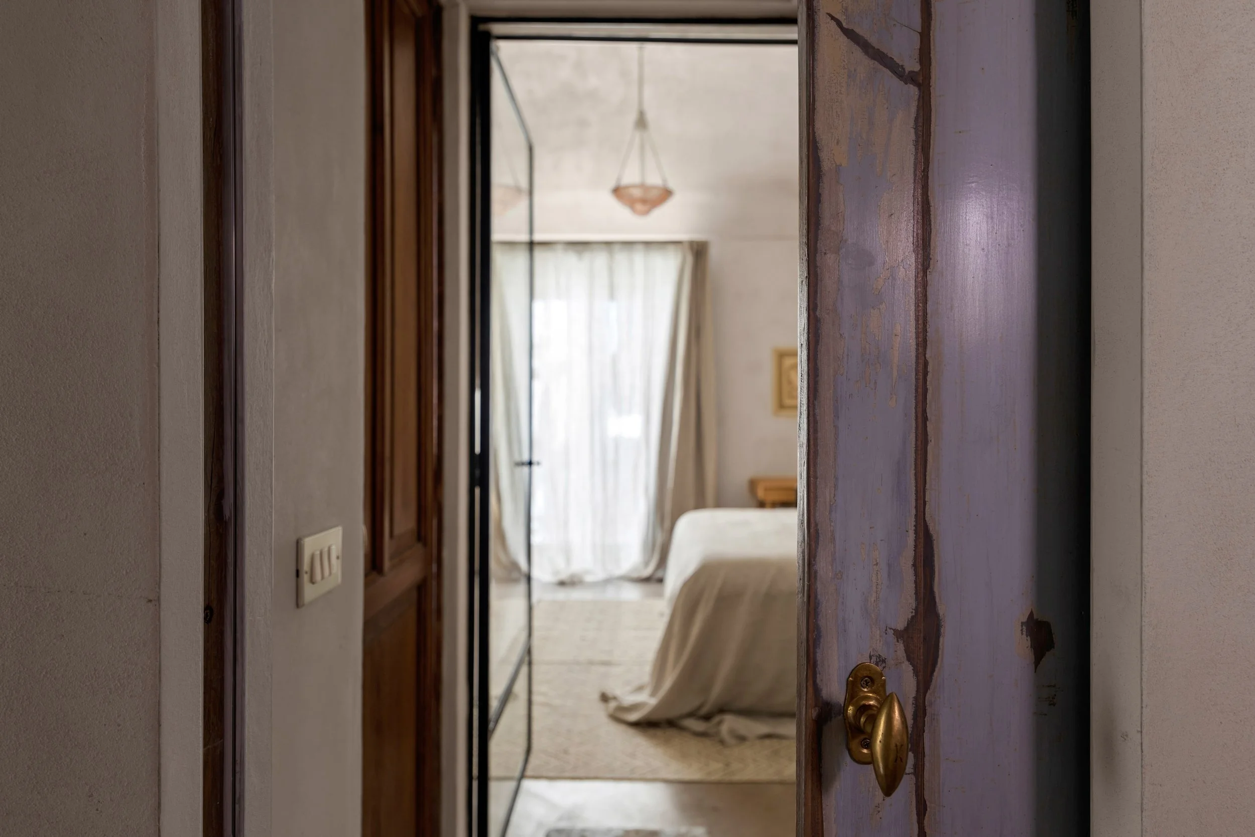 View through a doorway into a bedroom with a large bed, beige bedding, a window with curtains, and a hanging light fixture.