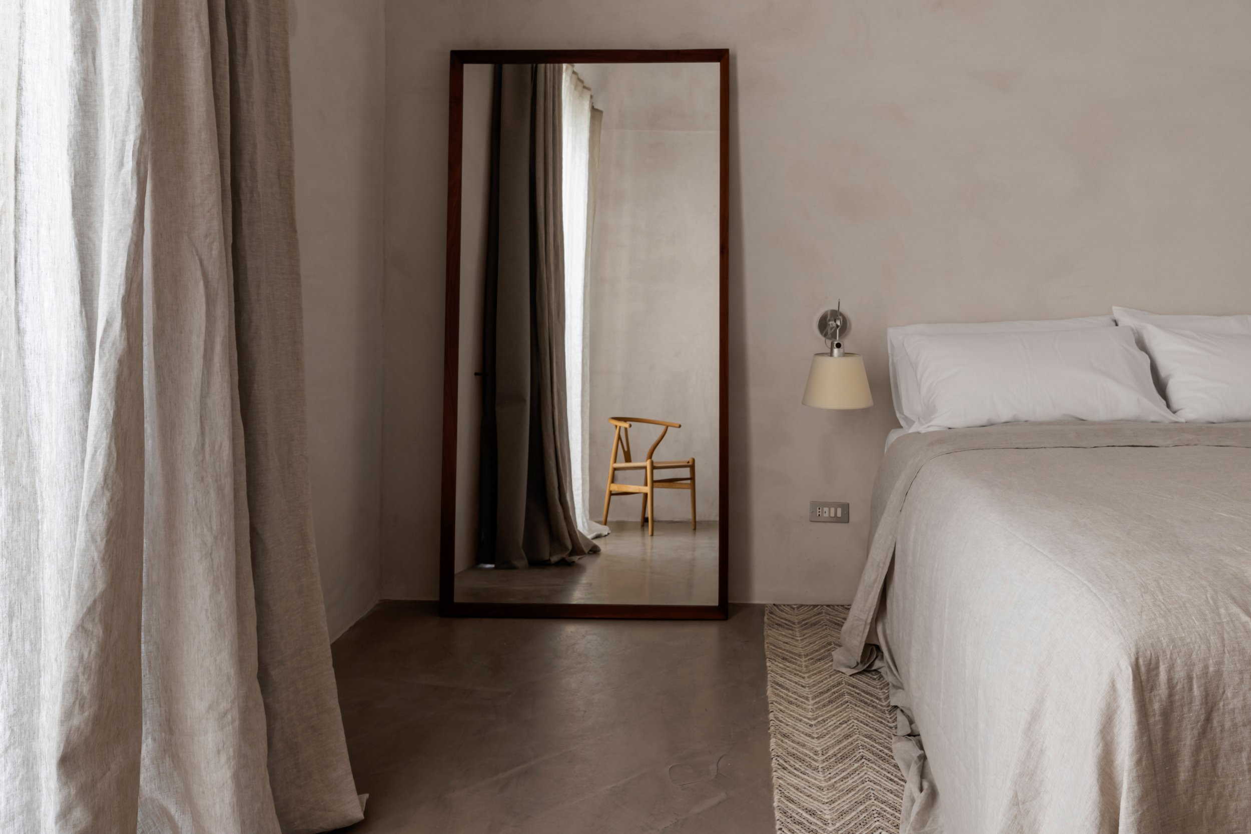 A bedroom with a bed, a wall-mounted lamp, a large mirror, and a wooden chair reflected in the mirror.