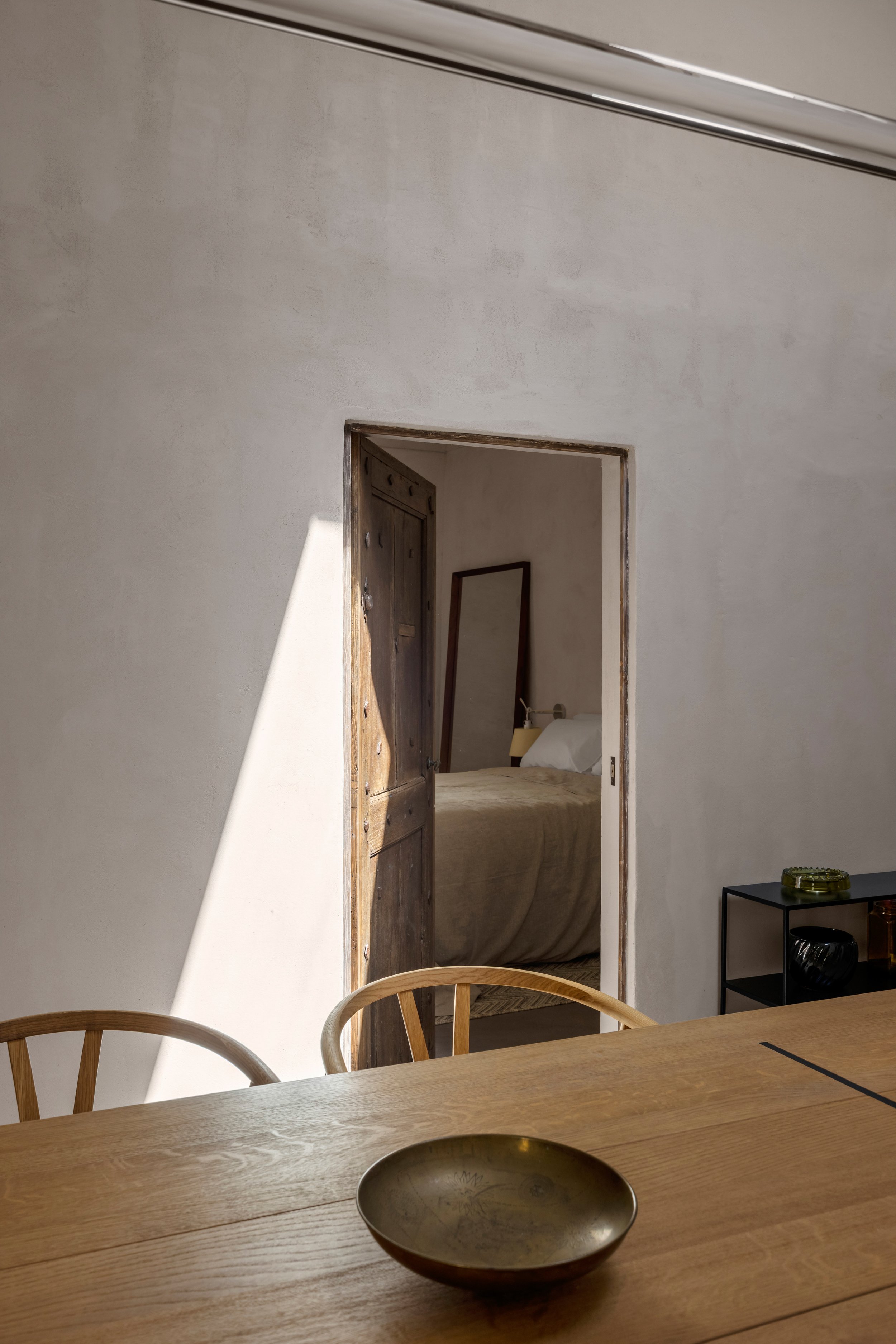 View through an open door into a bedroom with a bed, a mirror, and a beige side table.