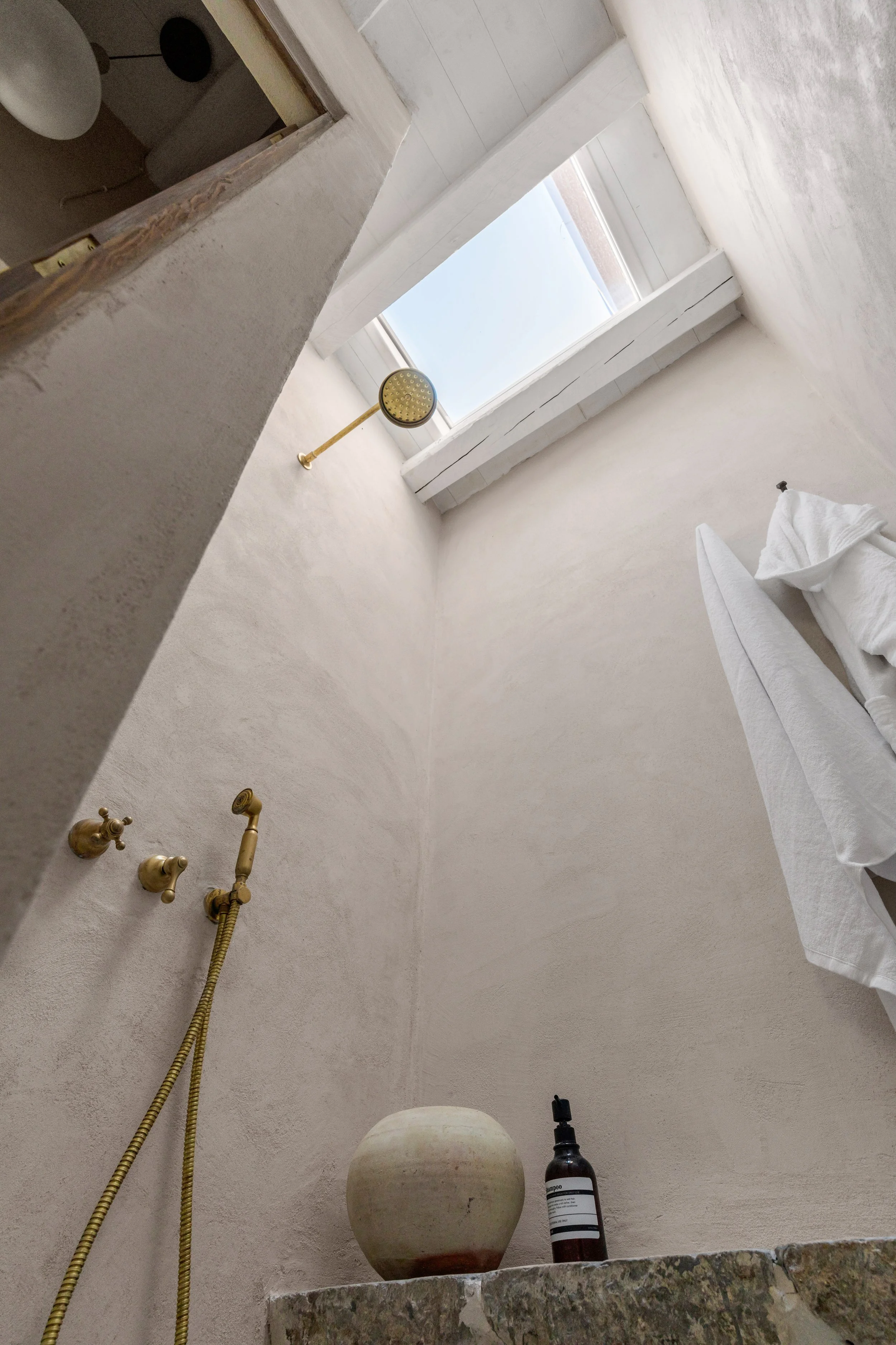 Interior view of a minimalist shower area with a beige textured wall, a gold showerhead, a beige ceramic vessel, and a soap dispenser, with a small window allowing natural light to enter.