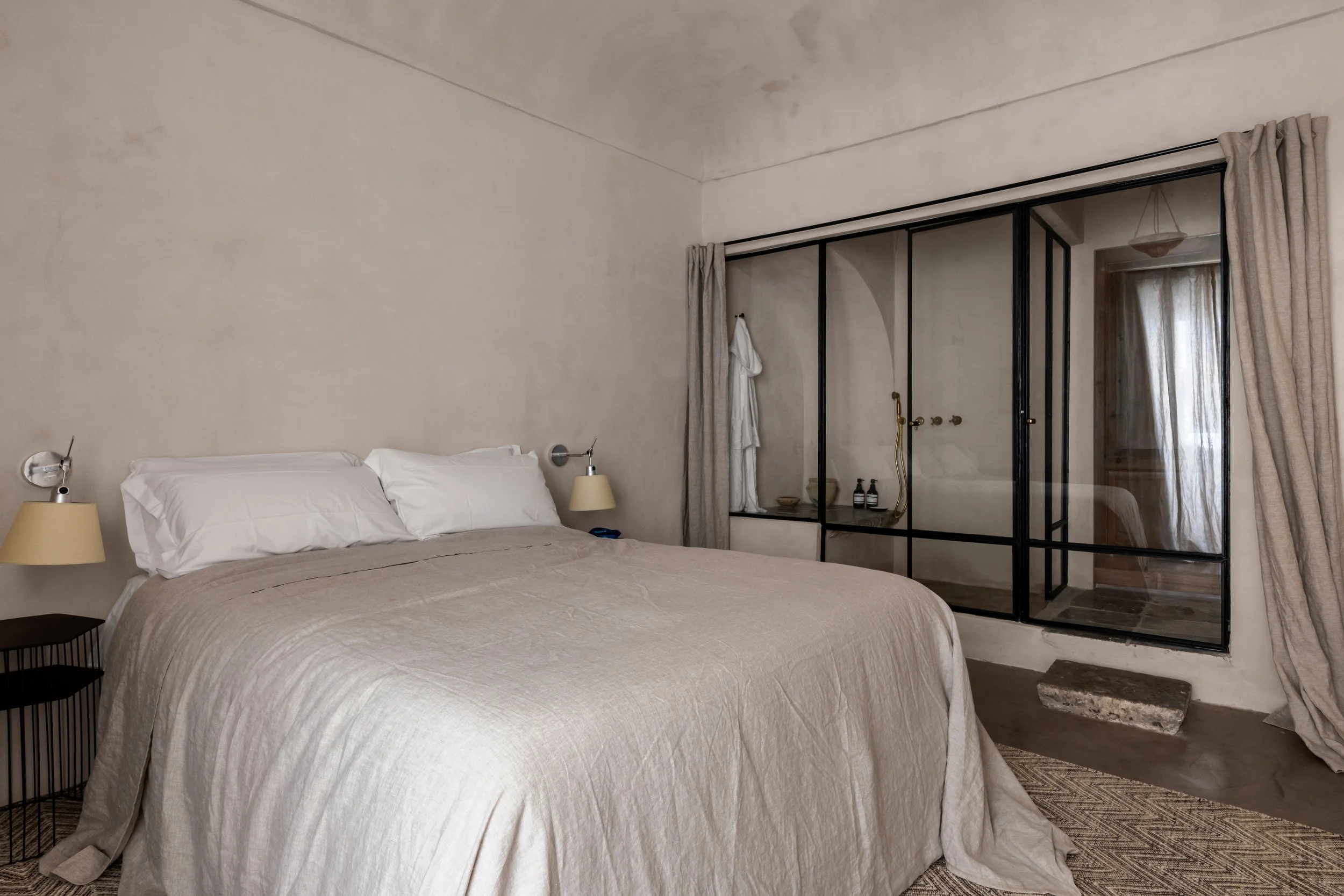 A bedroom with a minimalist design, featuring a large bed with white pillows and beige bedding, black nightstand, two wall-mounted reading lamps, and a glass shower enclosure with a black metal frame and beige curtains.