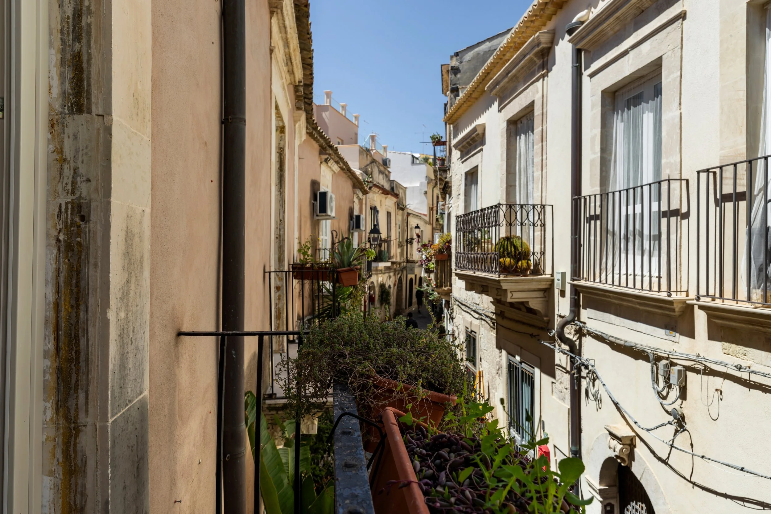 A narrow European street view with apartment buildings, balconies with potted plants, and laundry hanging outside under a clear blue sky.