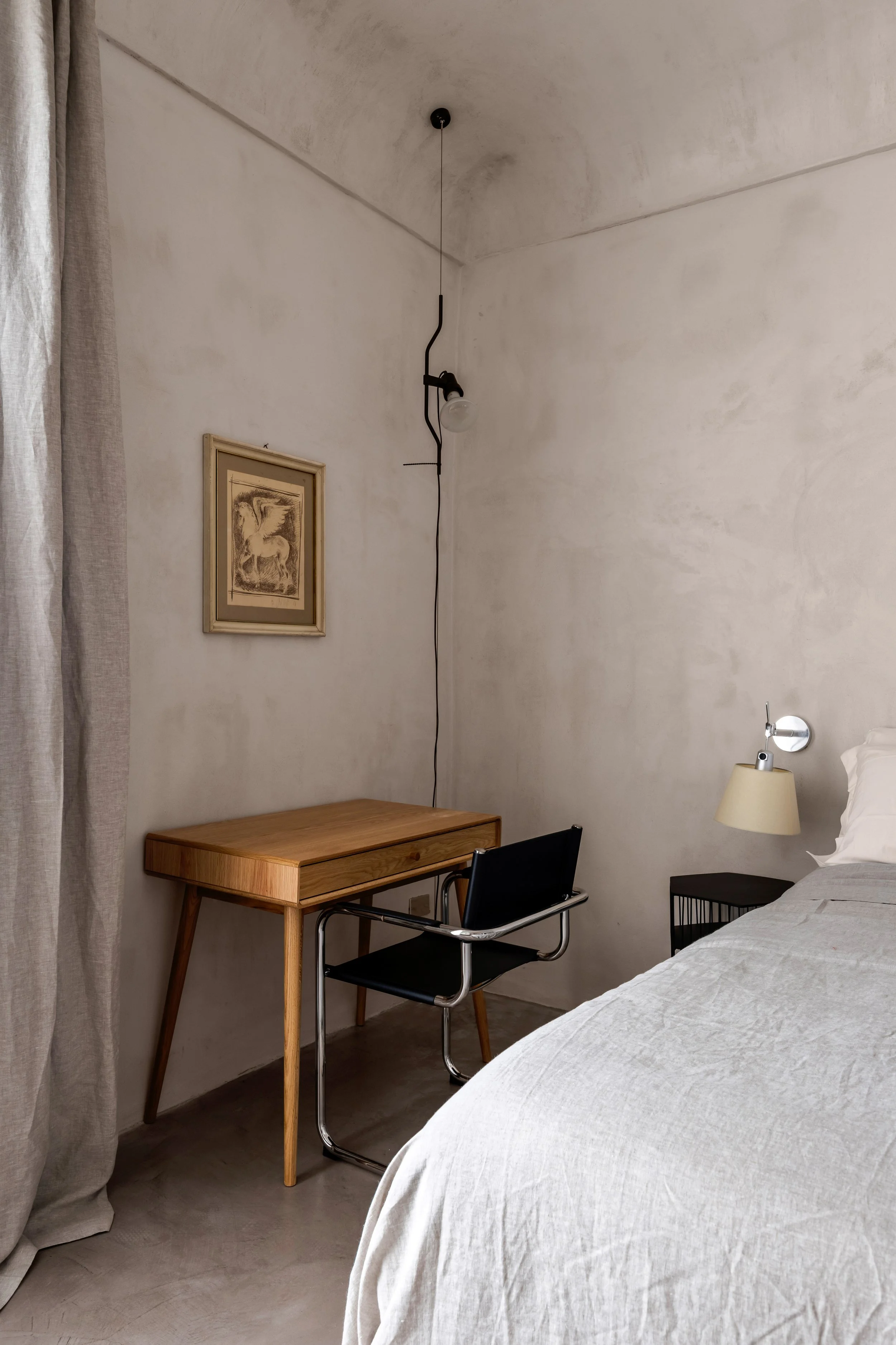 A minimalist bedroom corner with a wooden desk, black chair, bed with white linens, wall-mounted lamp, framed artwork, and a textured wall.