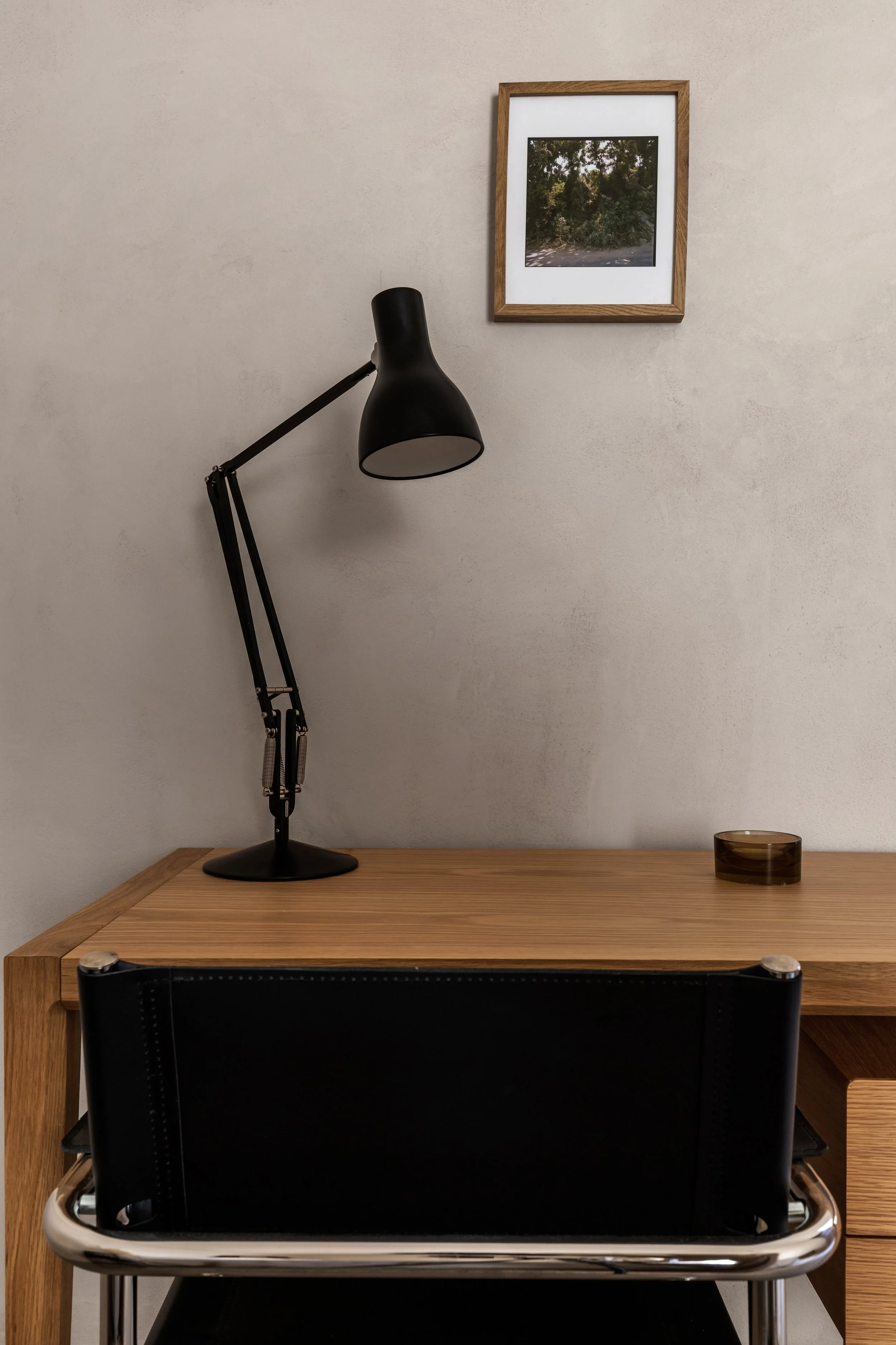 A minimalist office desk with a black adjustable lamp, a small brown glass container, and a framed picture of trees on the wall.