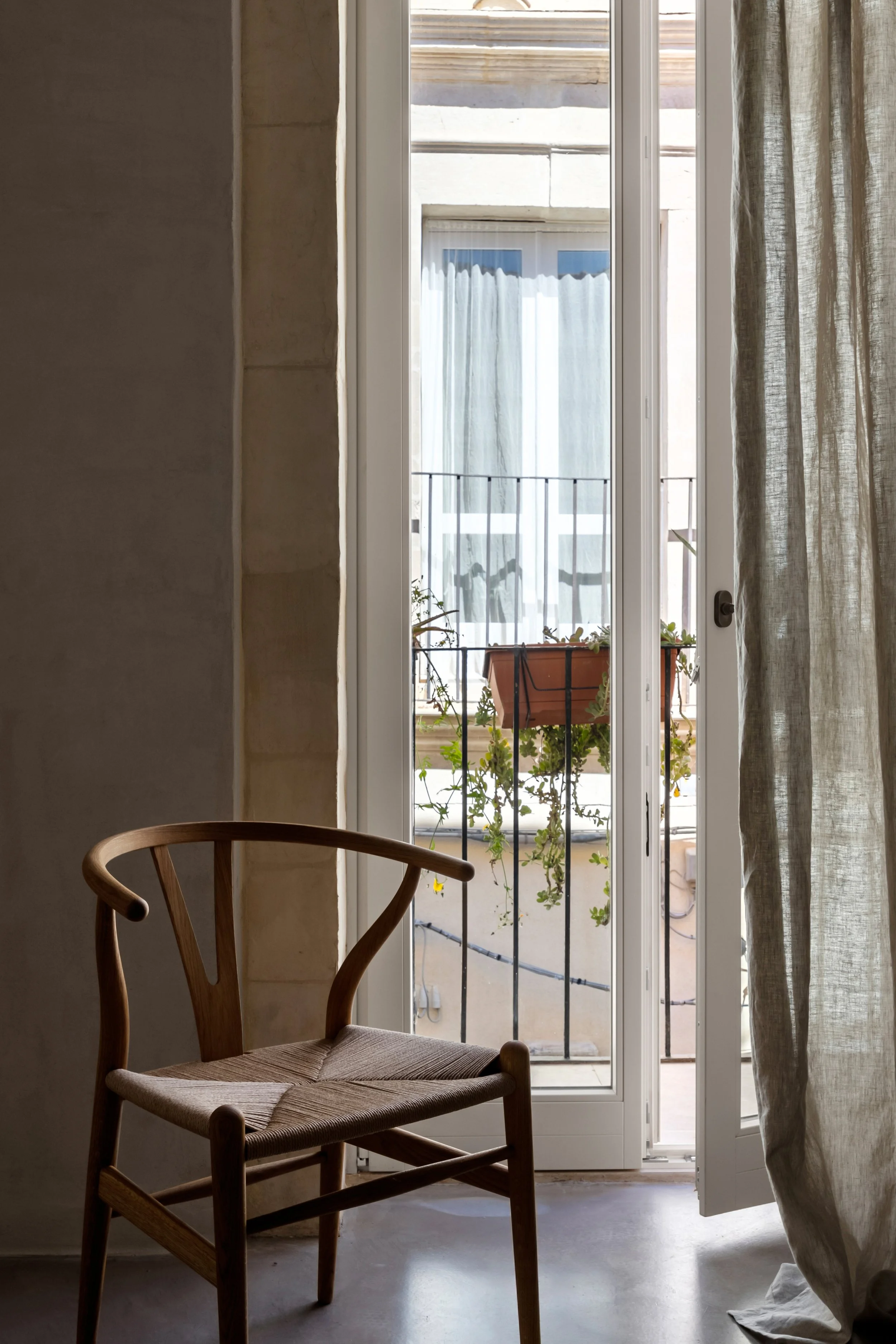 A wooden chair near a glass door with curtains, open to a small balcony with potted plants and a building facade in the background.