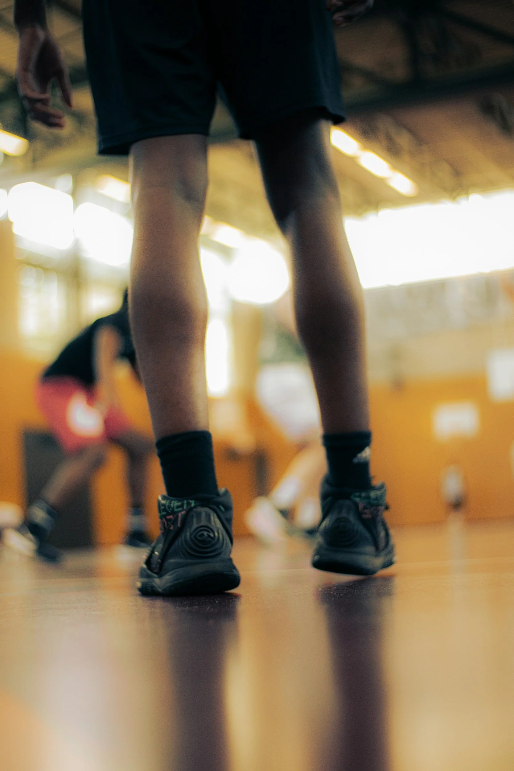 Close-up of sports shoes on a wooden gym floor with a person in athletic shorts in the background, in a sports or gym setting.