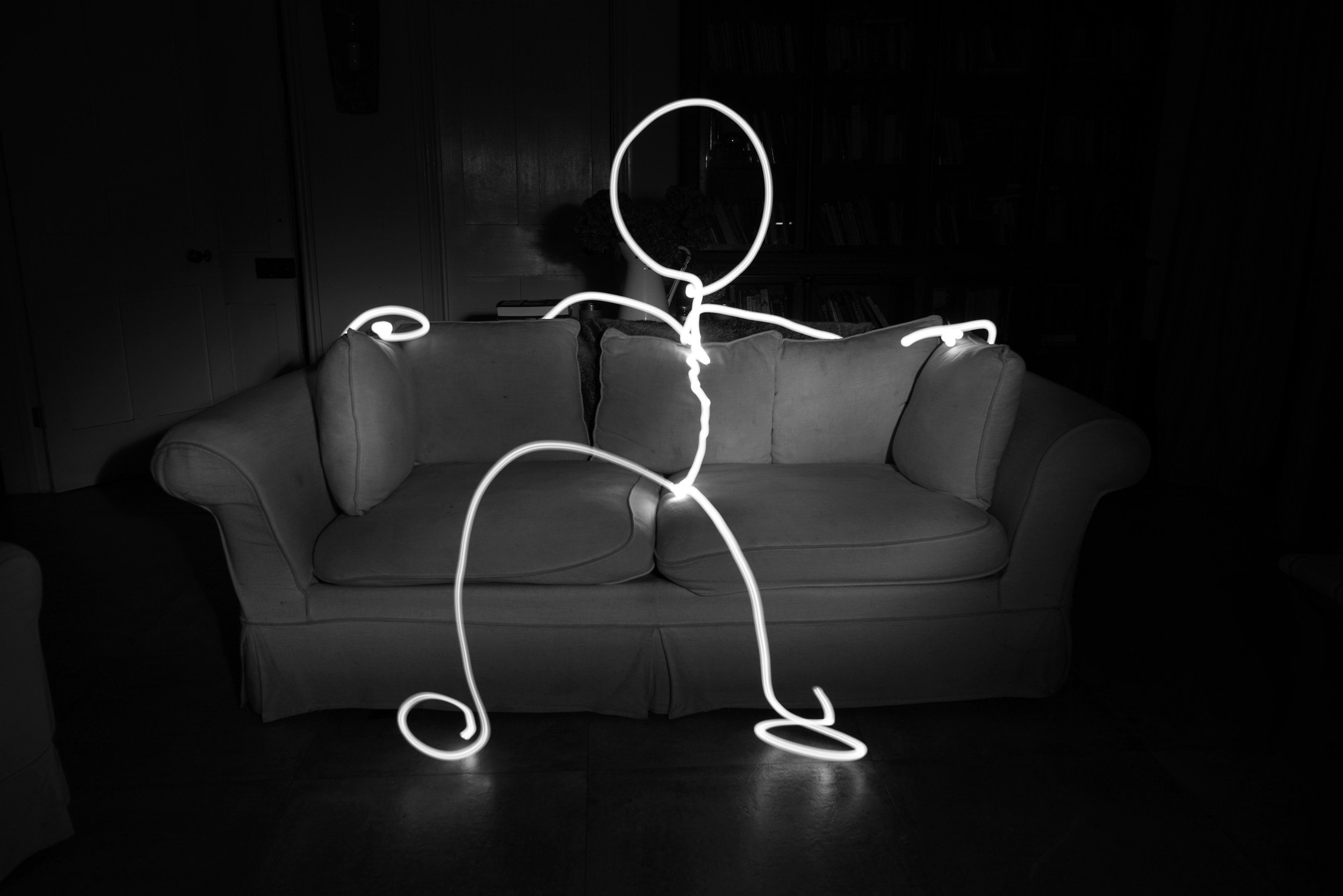 A neon light sculpture in the shape of a person sitting on a couch, positioned in a dimly lit room. The sculpture has an oval head and flexible limbs, with the wire outlines illuminated.