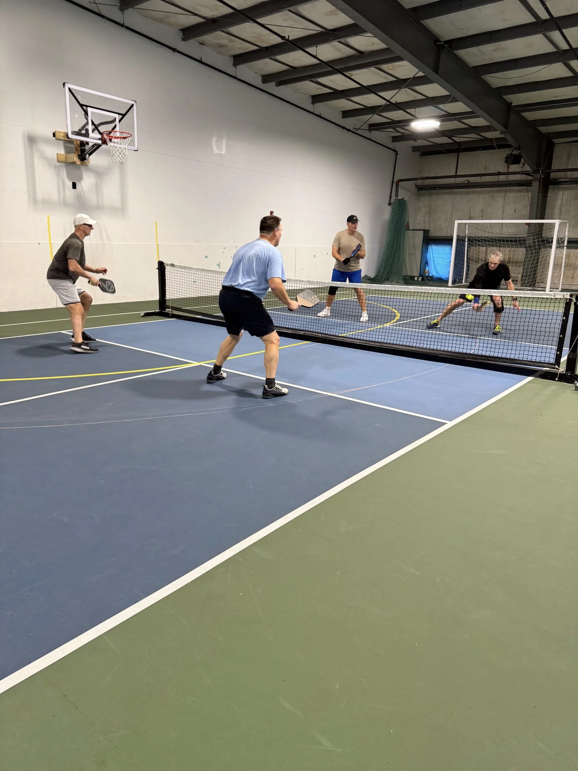 Four men playing pickleball indoors with a net, basketball hoop, and gym equipment in the background.
