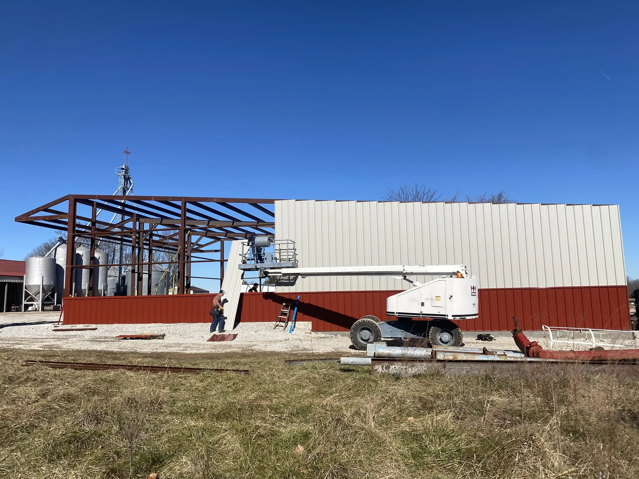 Construction site with workers, metal framework, and a white building under construction, with a blue sky in the background.