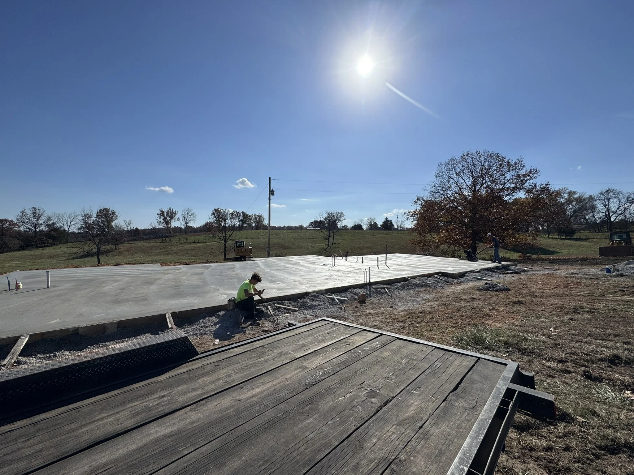 Construction site with workers pouring and smoothing concrete on a large slab outdoors, trees and a bright sun in the sky.