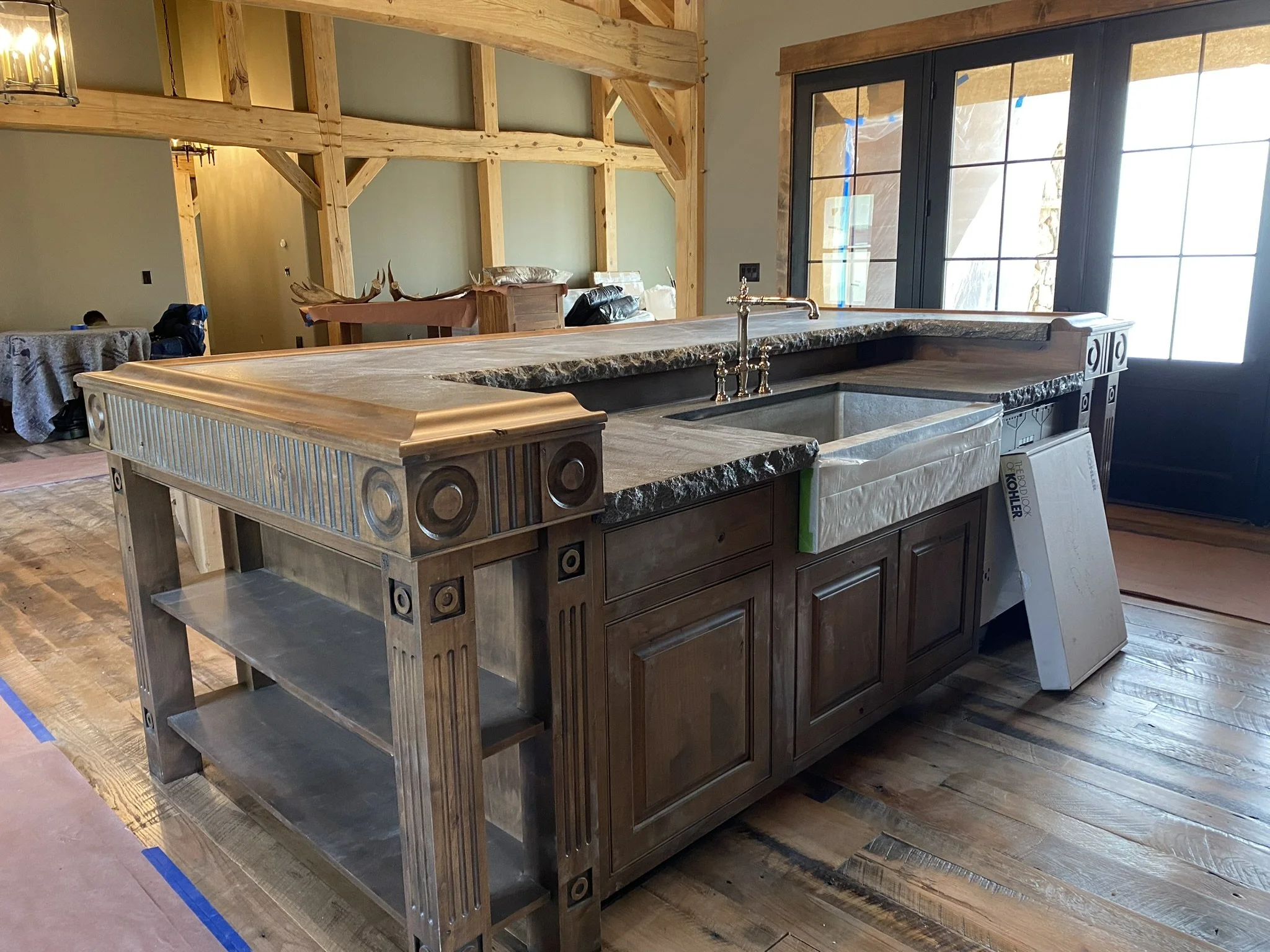 Interior view of a kitchen or bar area under construction, featuring a large wooden island with a marble countertop, a sink, cabinetry, and large windows with black frames letting in natural light.