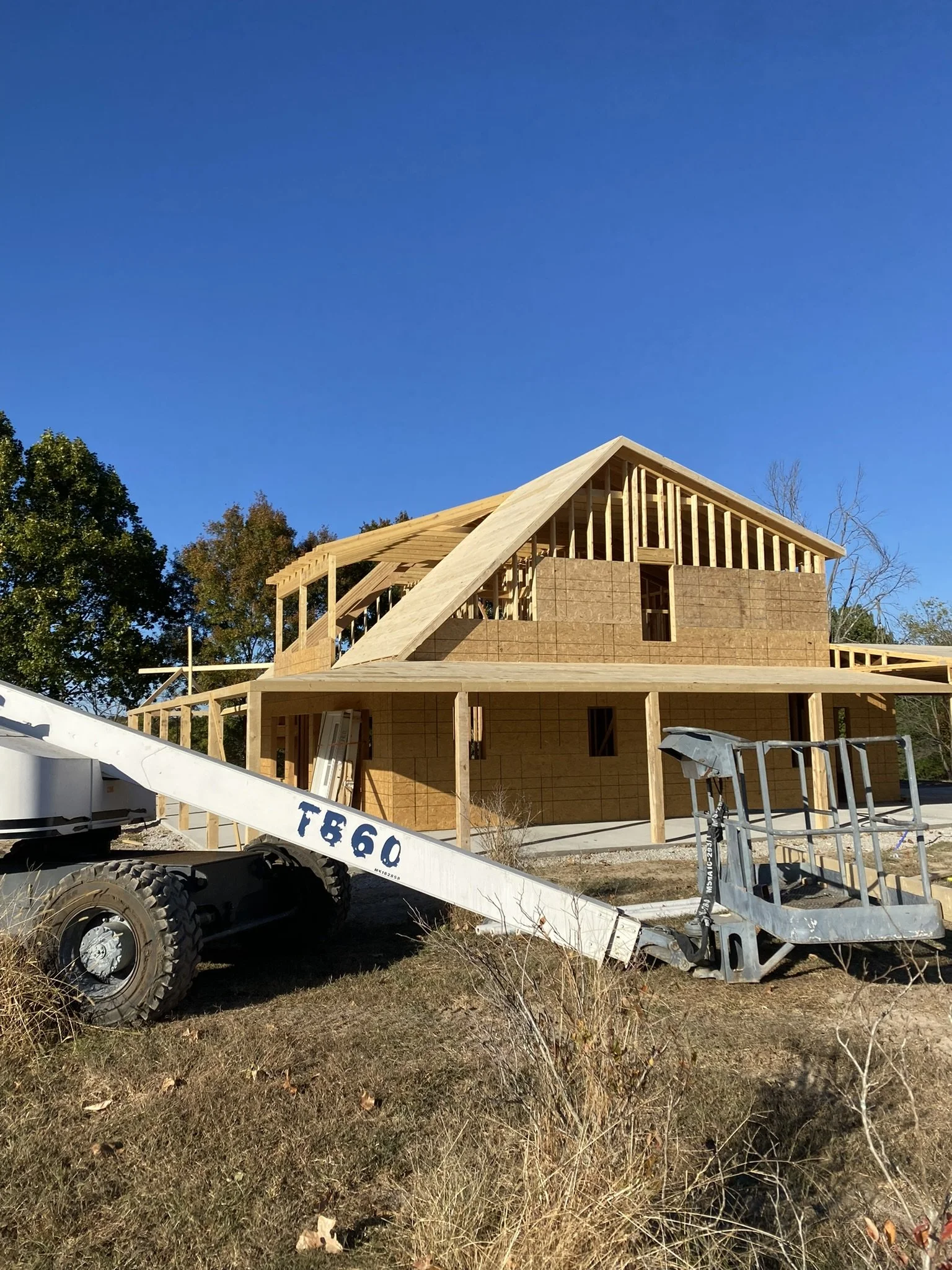 A house under construction with a wooden frame and a partially built brick wall. Construction equipment, including a mobile lift and scaffolding, are in the foreground. The sky is clear and blue with some trees in the background.