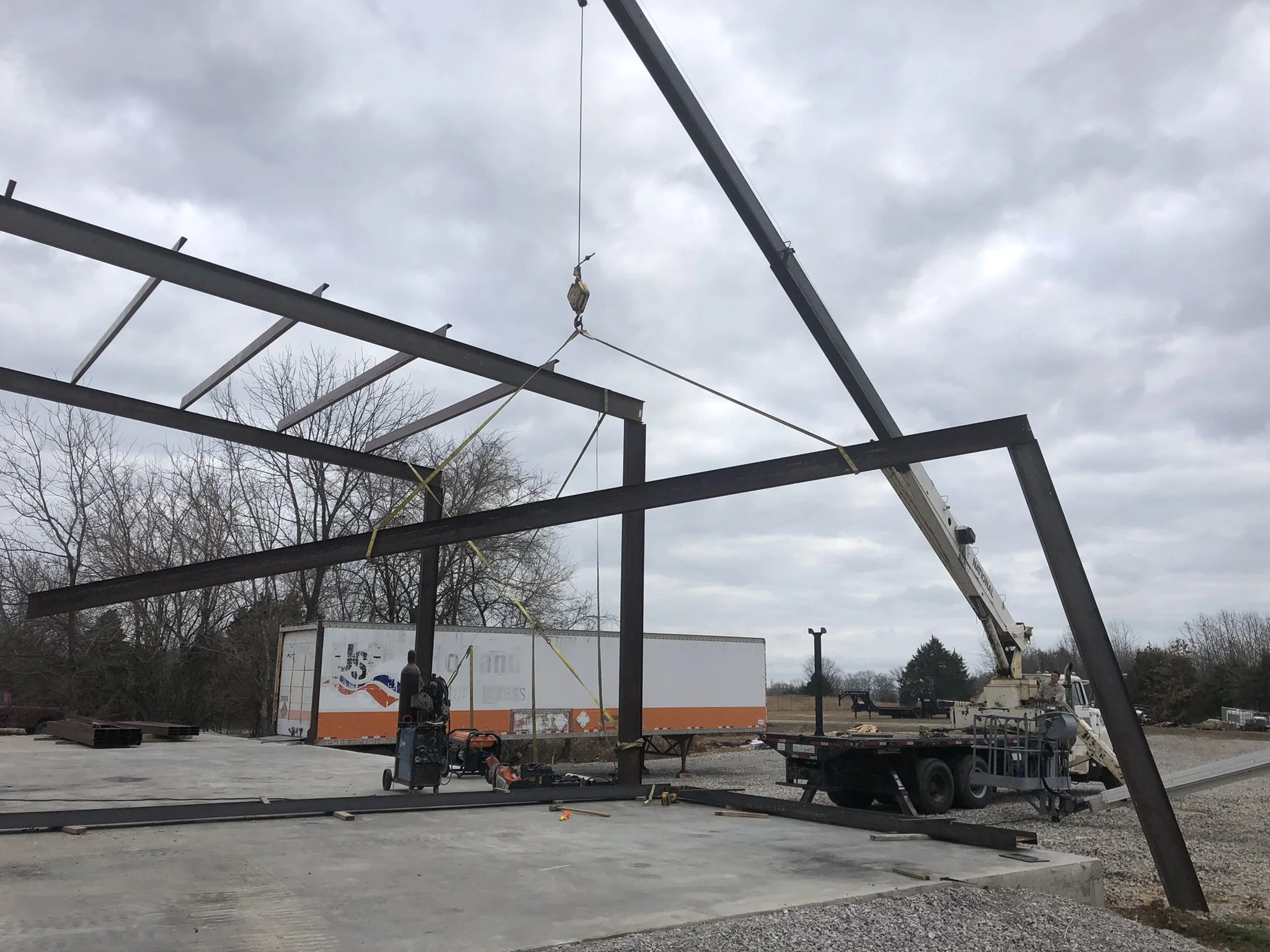 Construction site with steel framework, a crane lifting materials, and a large truck in the background, under cloudy skies.