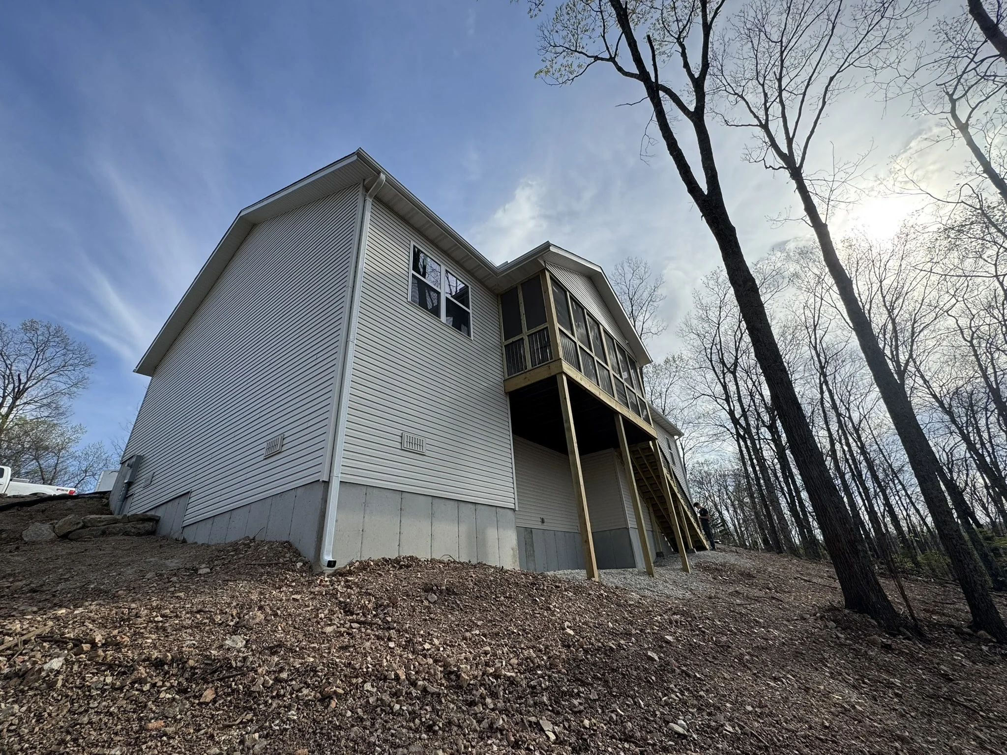 A multi-story house with light-colored siding and large windows, situated on a slightly elevated rocky terrain surrounded by tall, leafless trees under a partly cloudy sky.