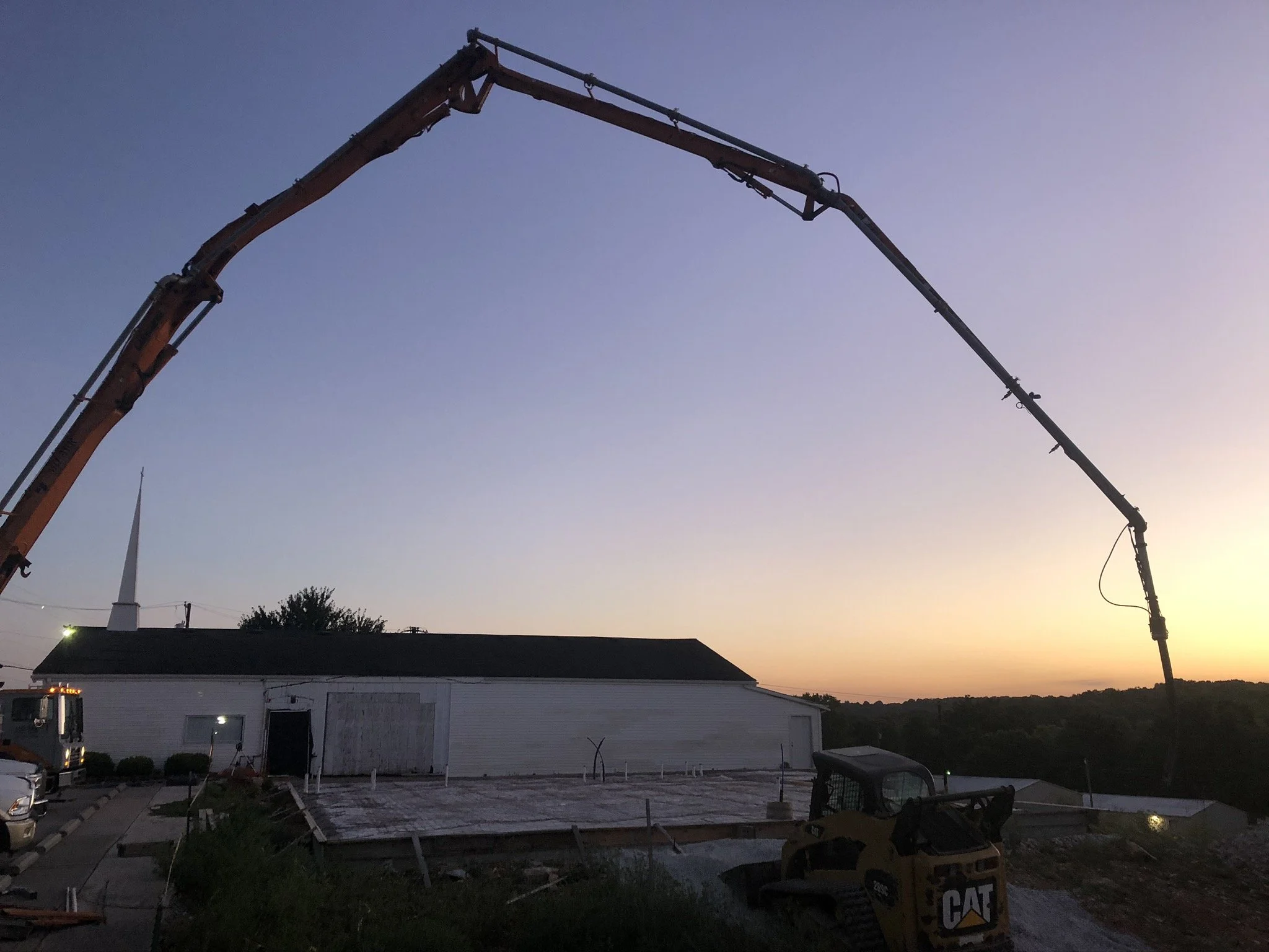 Construction site with a large crane extending into the sky during sunset. The crane is orange and appears to be used for building or demolition work. There is a white building in the foreground with a parking lot and some construction equipment, inc
