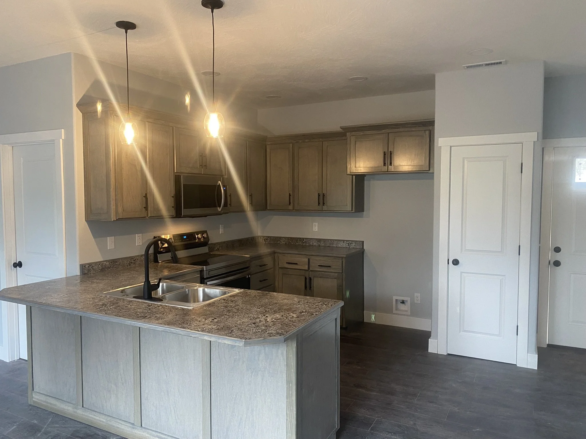 A modern kitchen with a large island featuring a granite countertop, a black kitchen sink and a high arching black faucet. The kitchen has light wood cabinets with black handles, a black microwave, and a stove. There are two hanging pendant lights ab