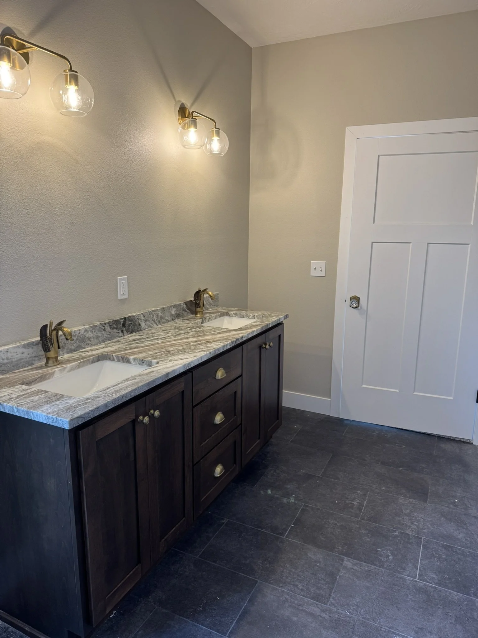 A bathroom vanity with a marble countertop, double sinks, and gold fixtures, set against beige painted walls with a white door and dark flooring.