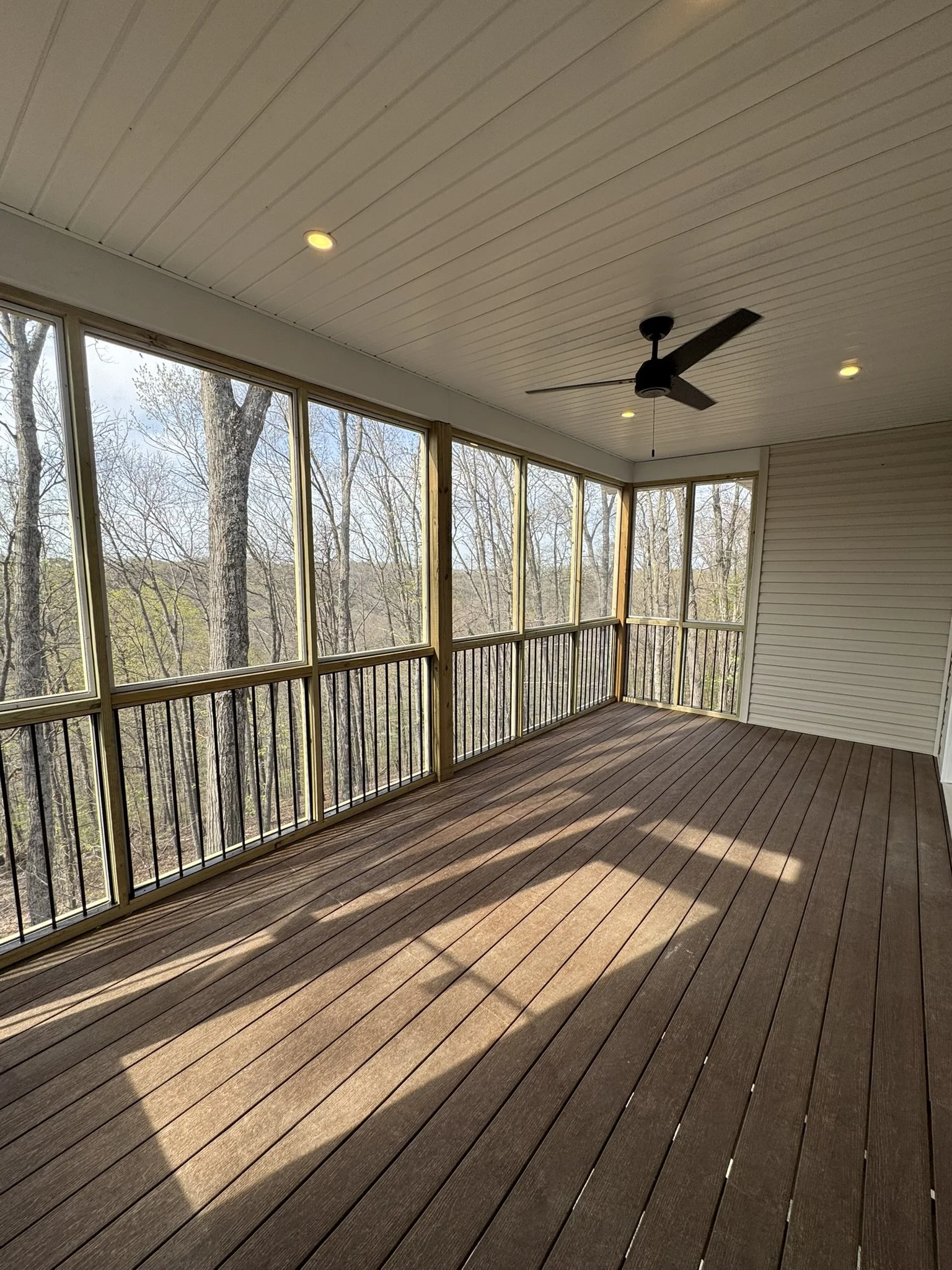 Enclosed porch with wooden flooring, ceiling fans, recessed lights, and large windows overlooking a wooded area.