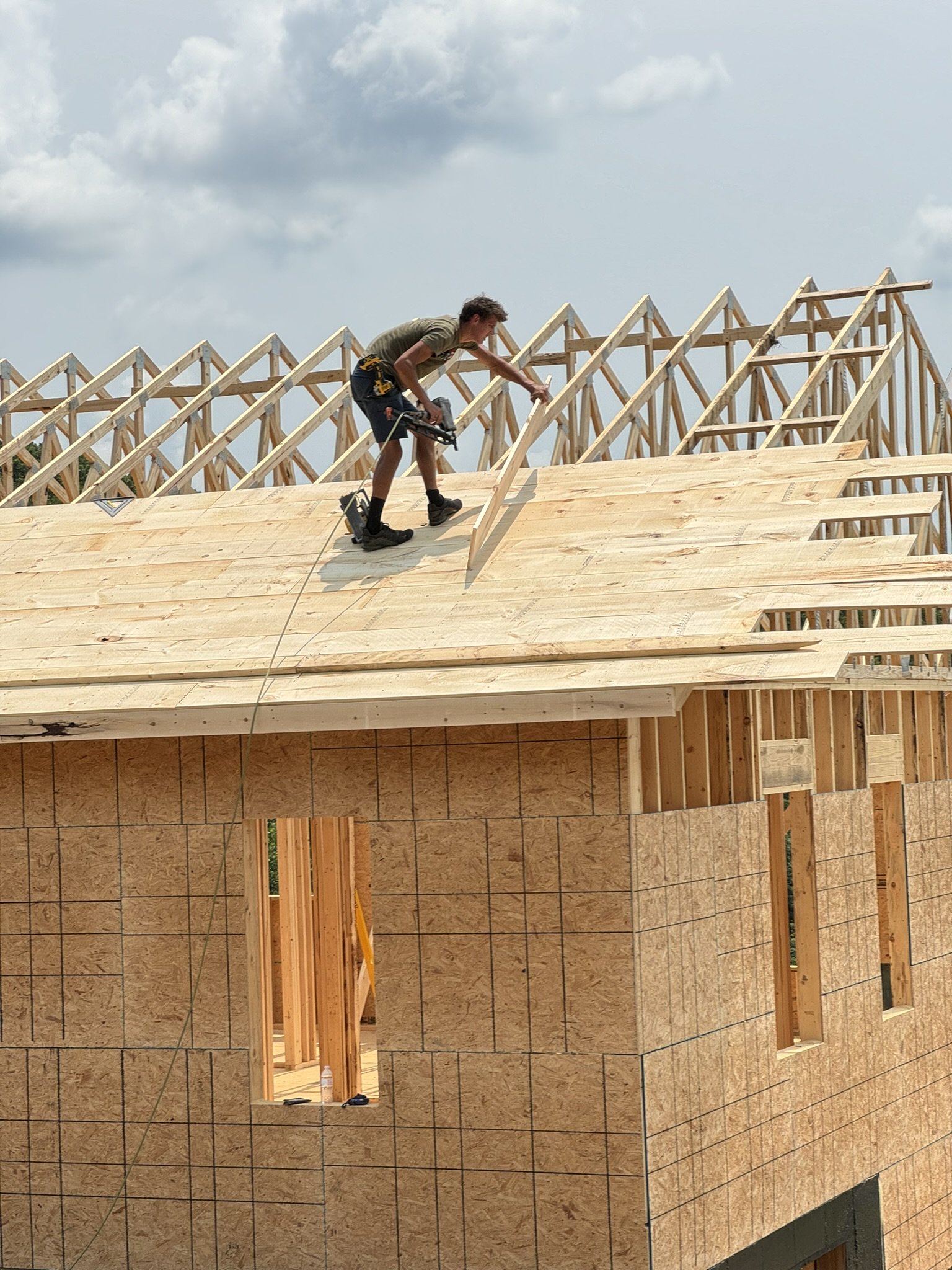 A man working on the roof of a house under construction, with unfinished wooden framing and plywood panels, against a cloudy sky.