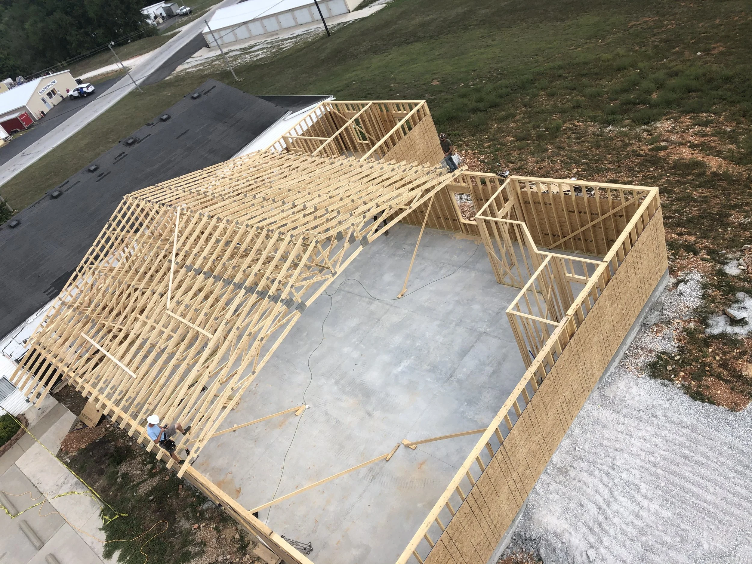 Construction site with a wooden framework for a building, two workers, one wearing a light blue shirt and hat, standing on the structure, and another wearing dark clothing and a safety helmet. The foundation is concrete, and there is a grassy area wi