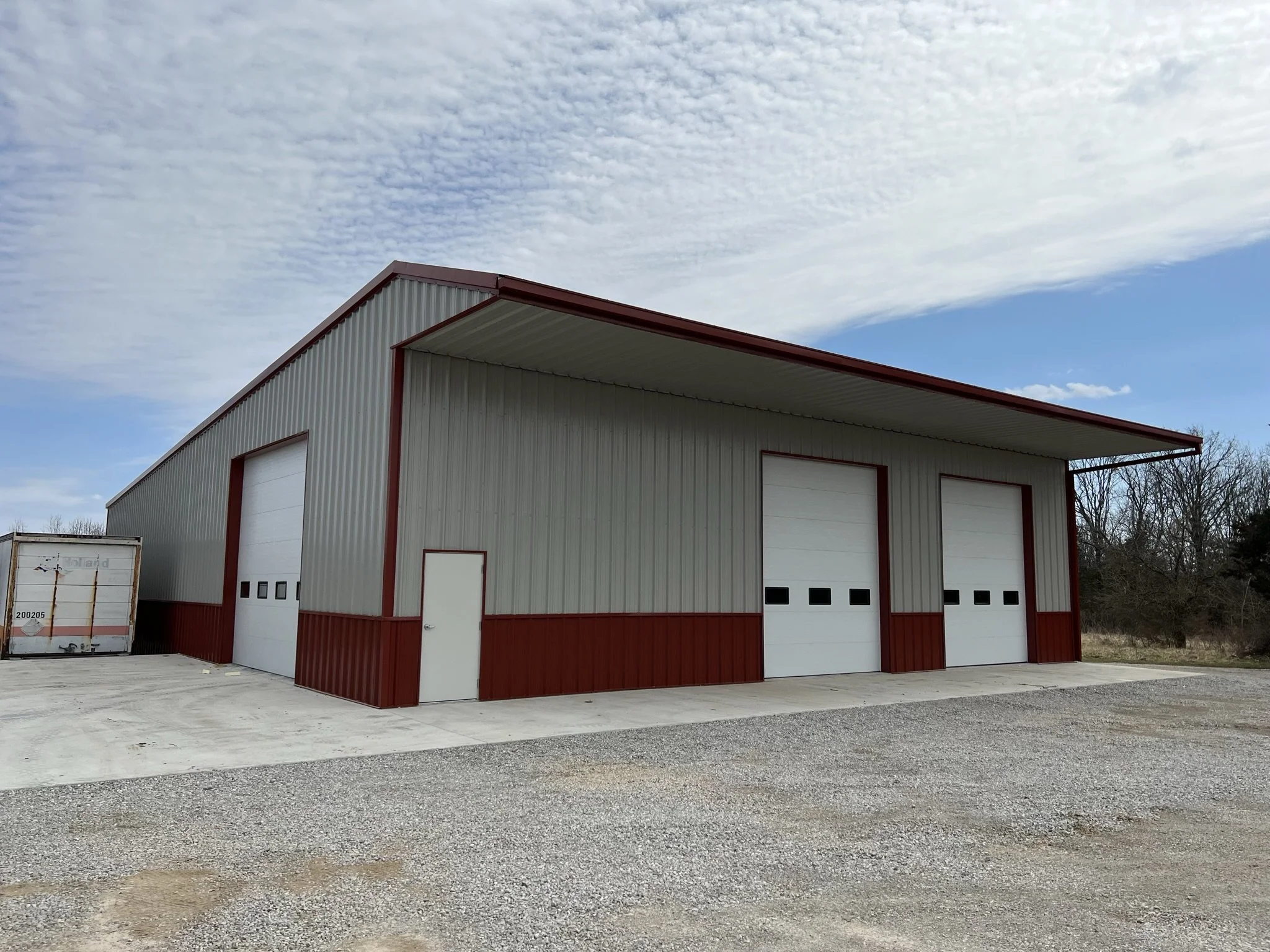 Large metal building with red trim and three garage doors, a small white door, and a gravel parking area in front, under a partly cloudy sky.
