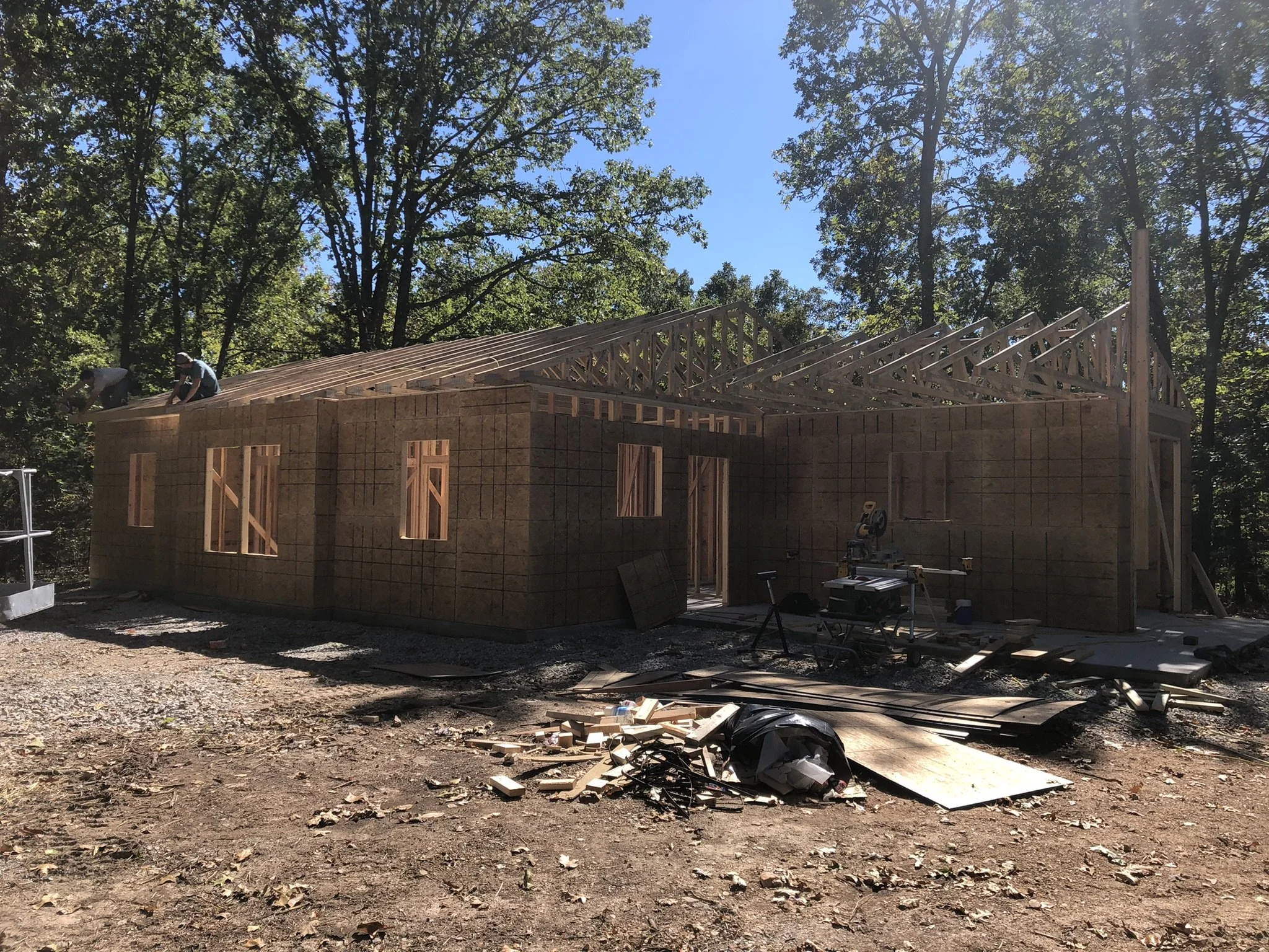 A house under construction with a wooden frame structure, workers on the roof, surrounded by trees and construction debris on the ground.