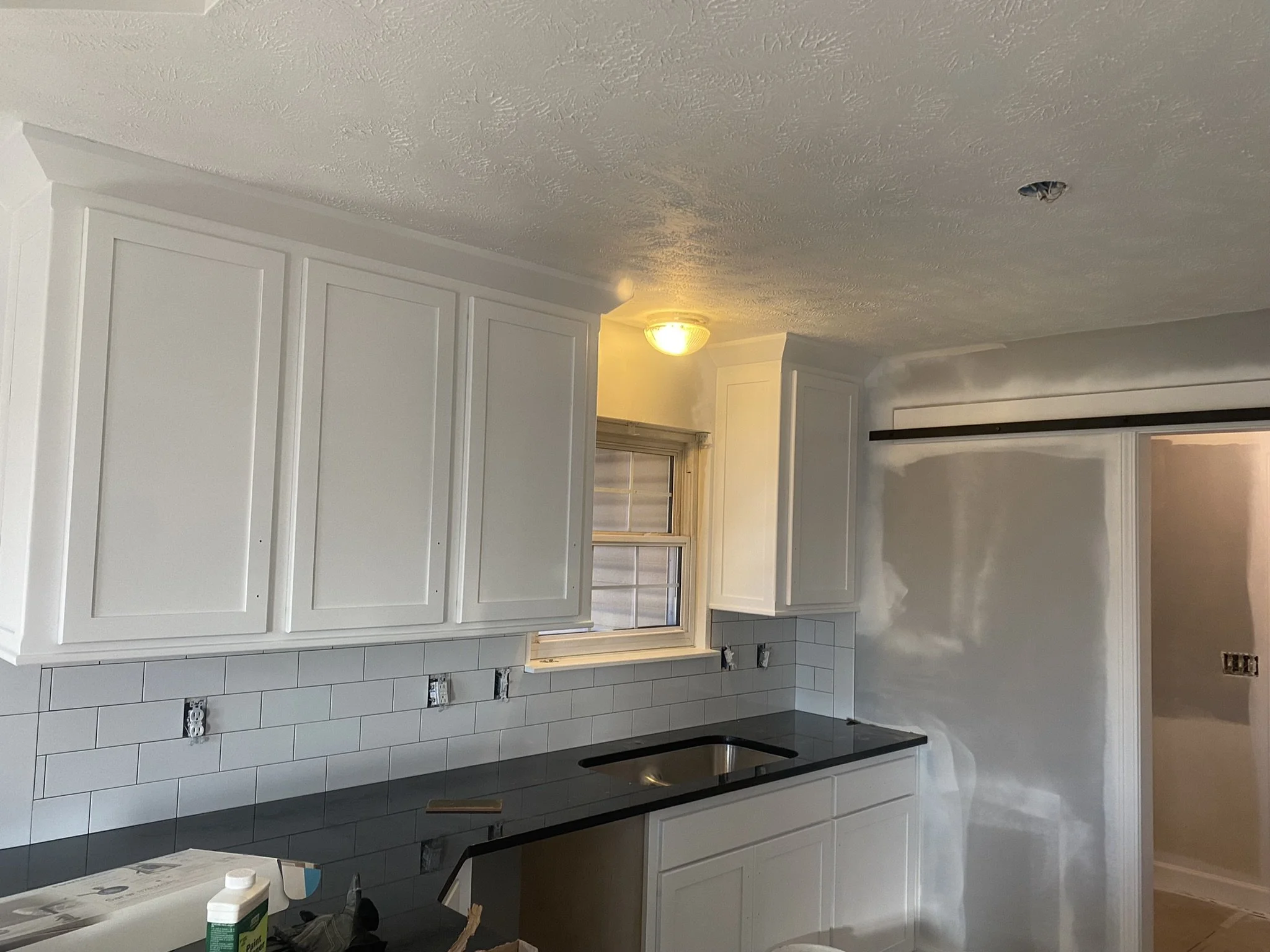 Kitchen with white cabinets, a window, and a black countertop. There is a light fixture on the ceiling and an electrical outlet with multiple sockets on the wall.