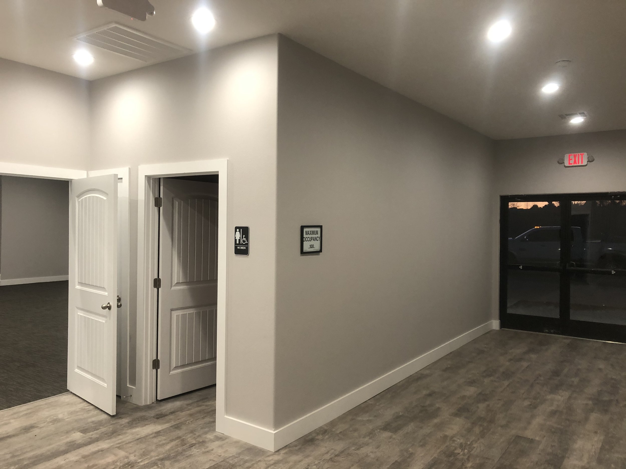 Interior of a building with a white wall, ceiling-mounted lights, an exit sign, a door with a handicap access sign, and a glass door showing a parking lot outside during dusk.