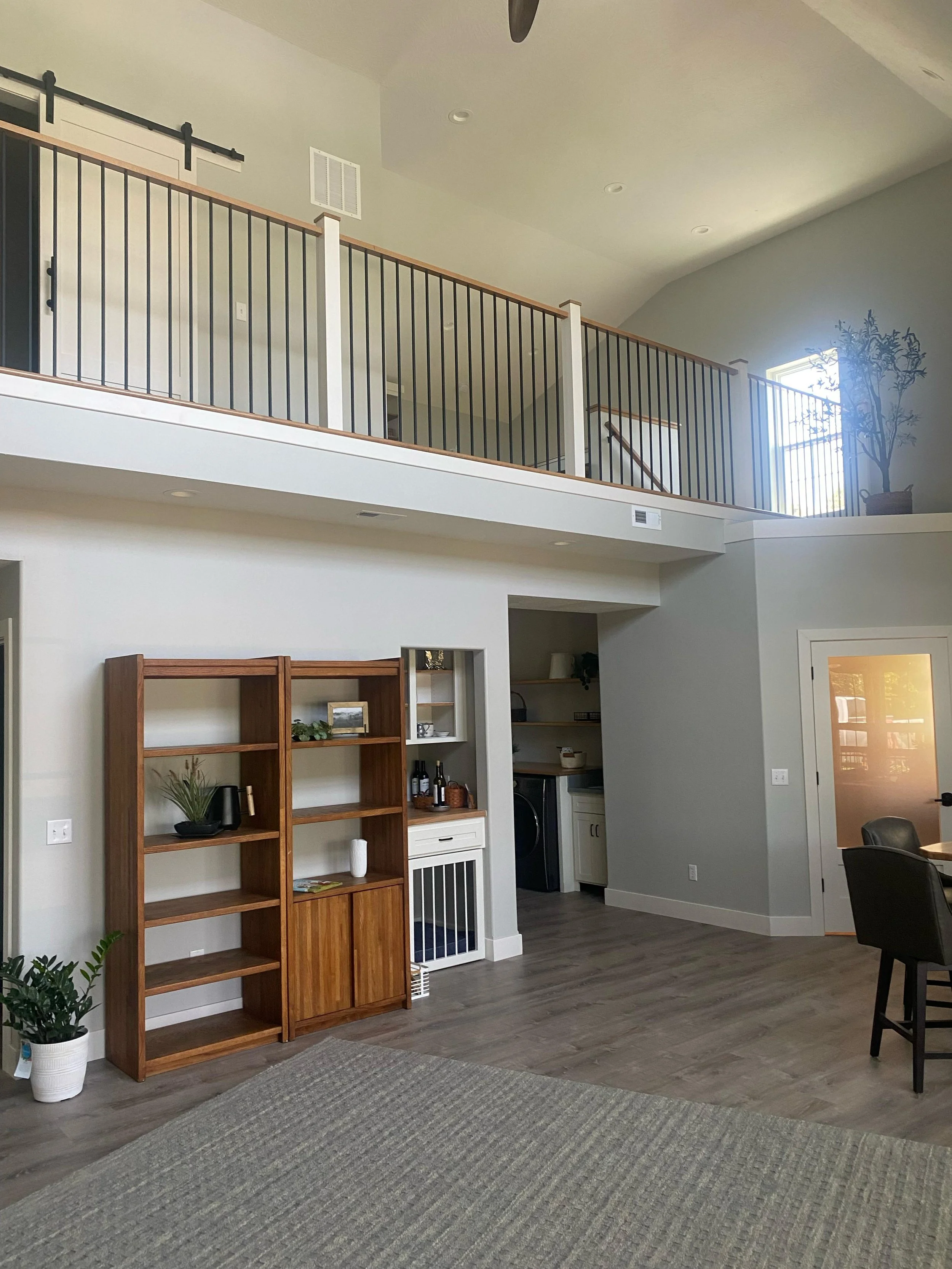 Living room with a wooden bookshelf, a potted plant, a gray textured rug, and an open door leading outside. A staircase with a black metal railing is visible on the upper level, and a large window allows natural light to fill the space.