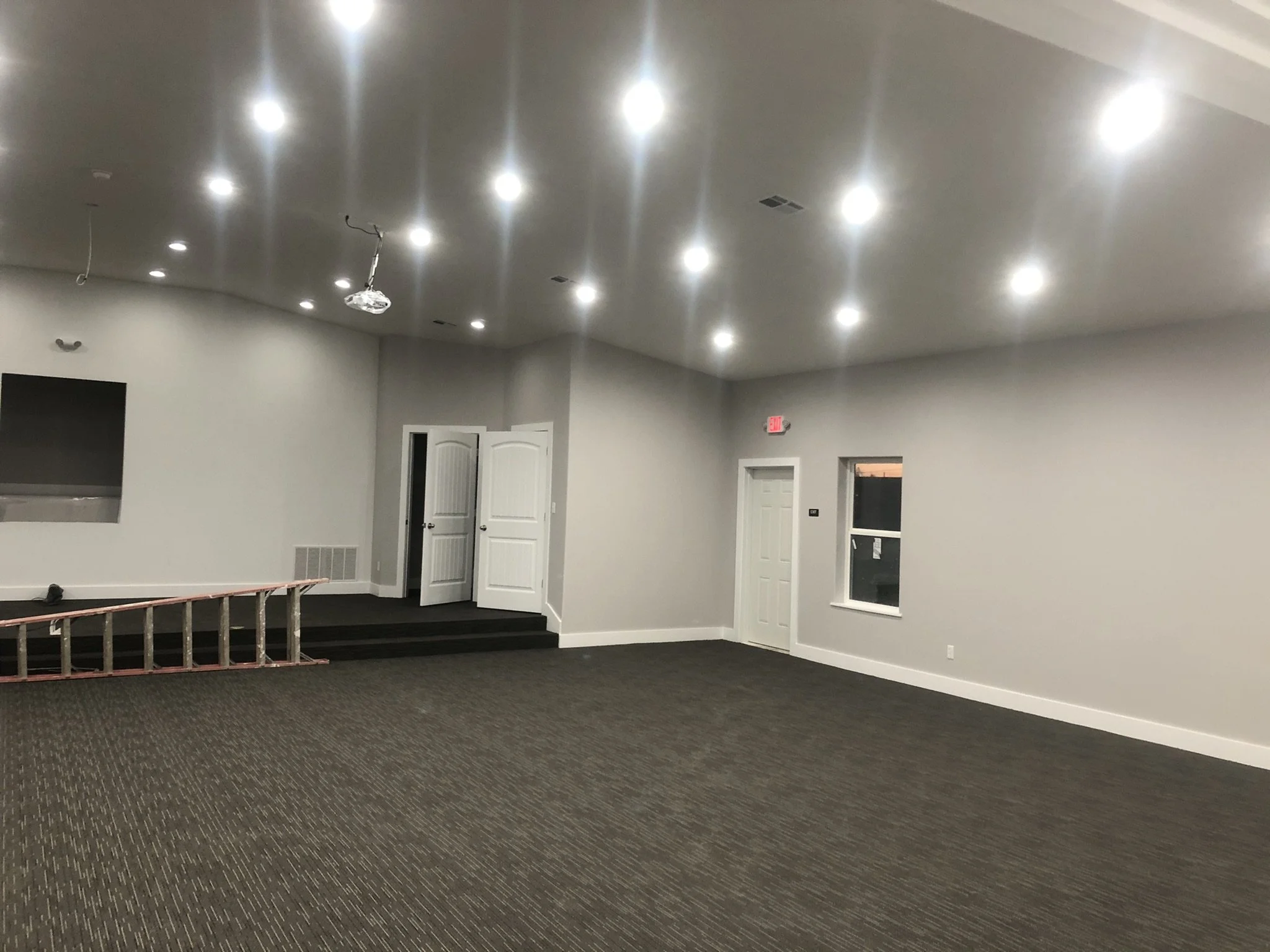 An empty room with several ceiling lights, two white doors, a window, and a carpeted floor. There are also some wires hanging from the ceiling and a ladder on the left side.