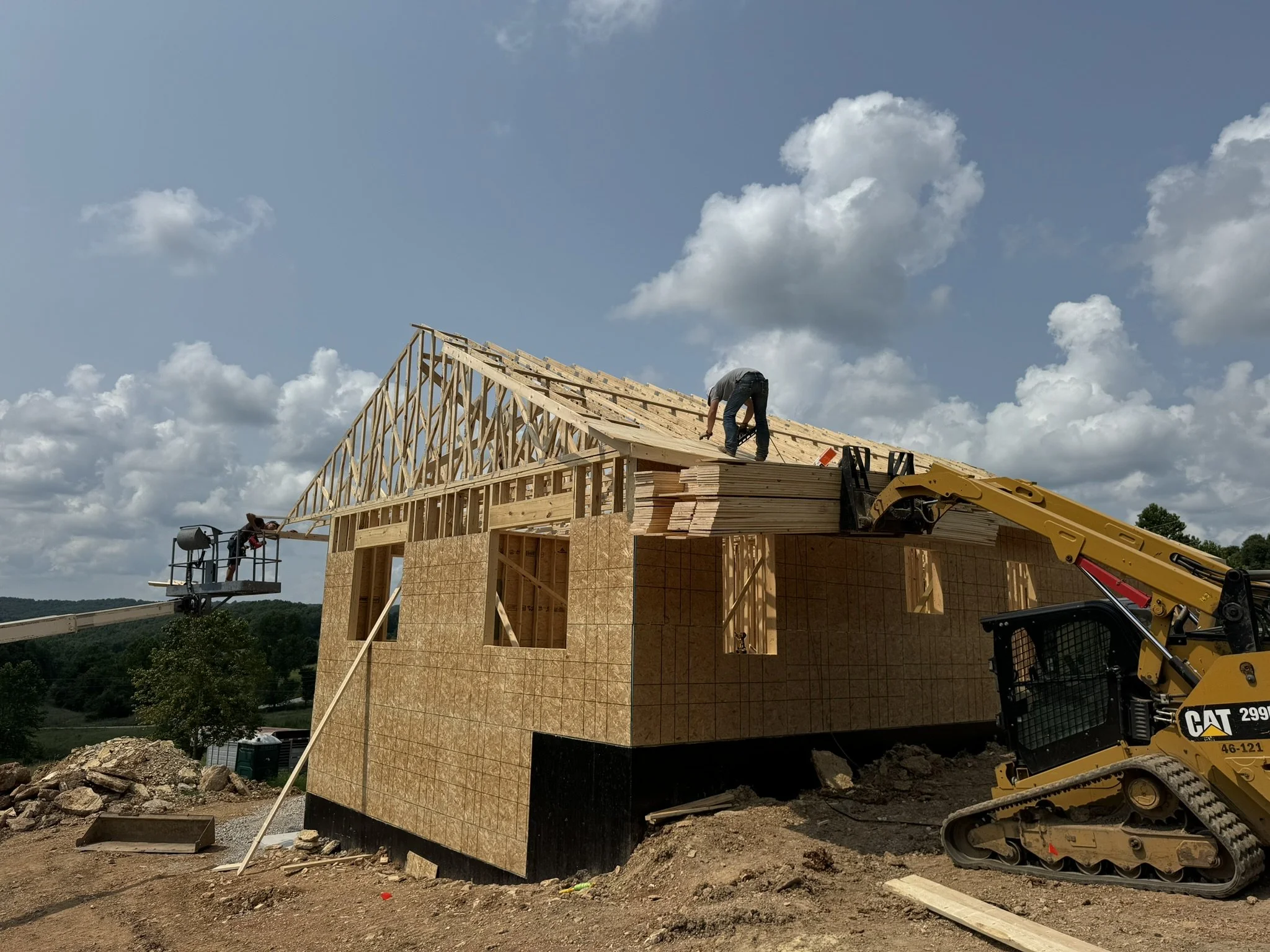 A construction worker installs roofing on a wooden house frame under a partly cloudy sky. A forklift is present on the right side of the image, and another worker is operating a lift on the left side.