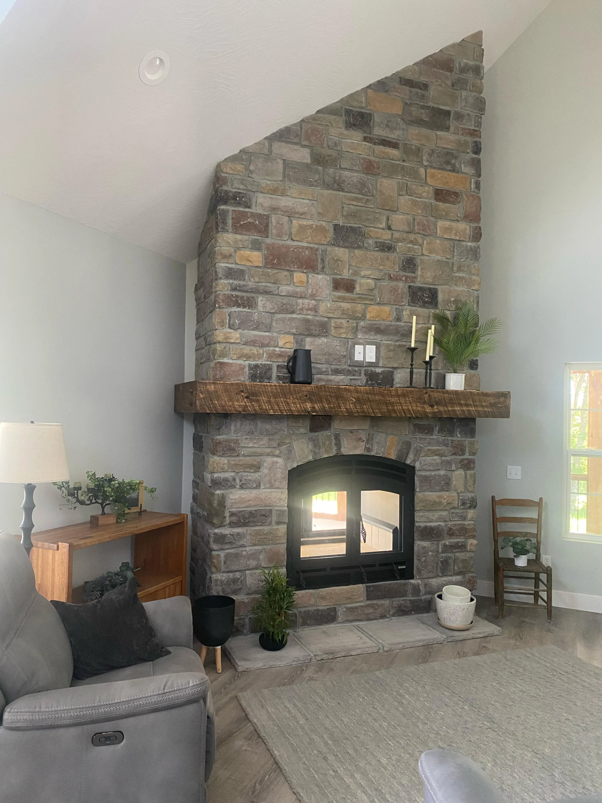 Interior of a living room with a brick fireplace, wooden mantle, and various decorative items including candles, a potted plant, and small plants. There is a gray couch with black pillows, a lamp, and a small wooden side table. The room features ligh