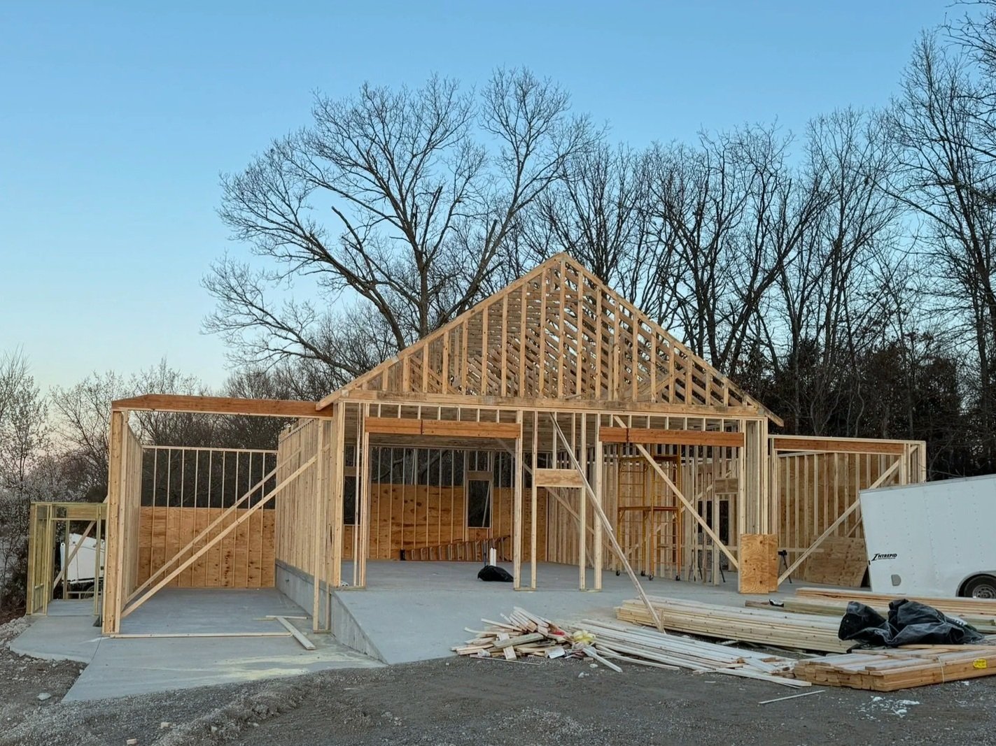 House under construction with wooden framing and a concrete foundation, surrounded by construction materials and equipment, with leafless trees in the background.