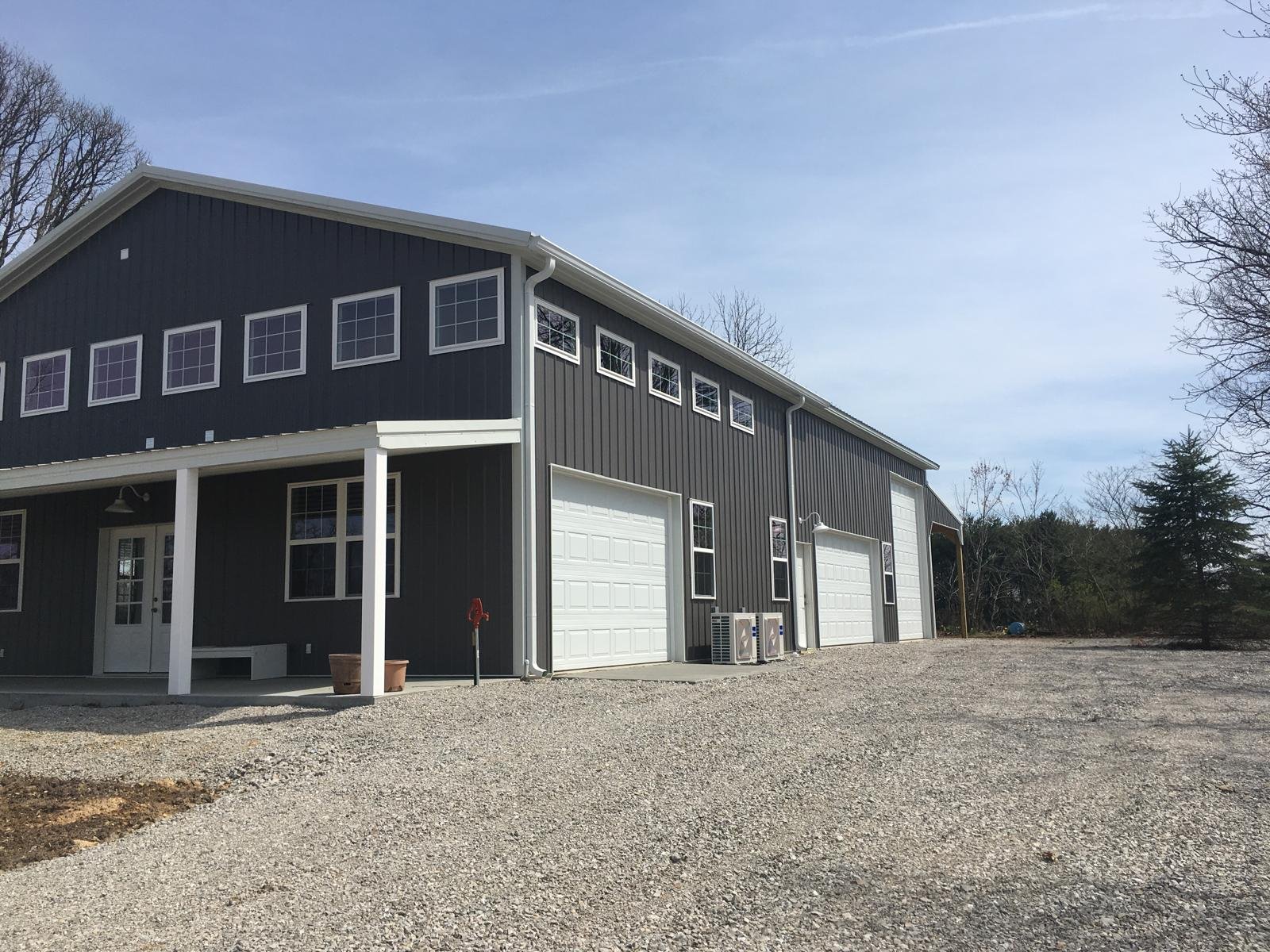 A large metal building with three garage doors, red trim, and a sloped roof, situated on a concrete and gravel lot under a partly cloudy sky.