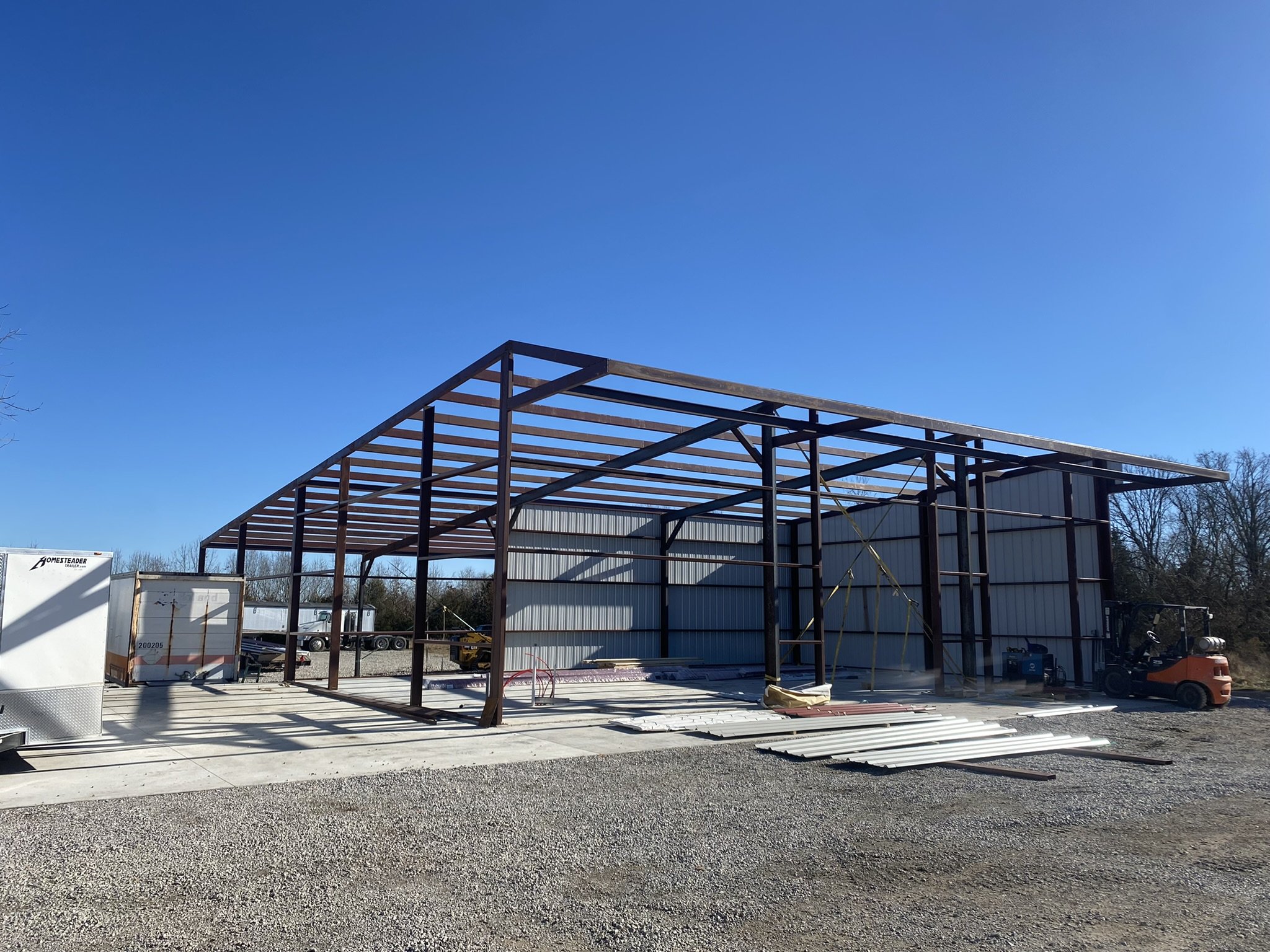 Construction site with a steel frame structure being built, surrounded by gravel ground, various construction materials, and equipment, under a clear blue sky.