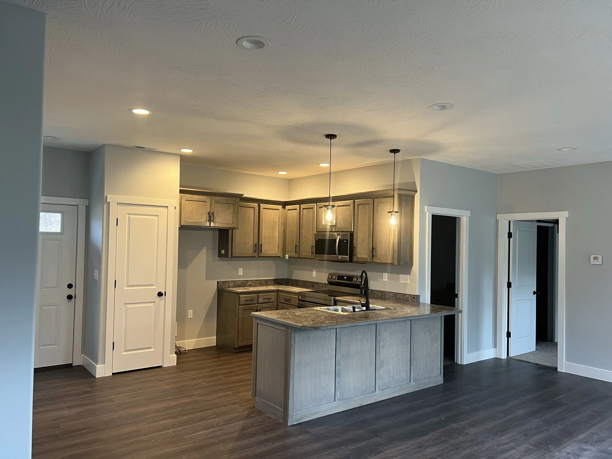 A modern kitchen with wooden cabinets, pendant lights over a kitchen island, stainless steel appliances, and dark wood flooring.