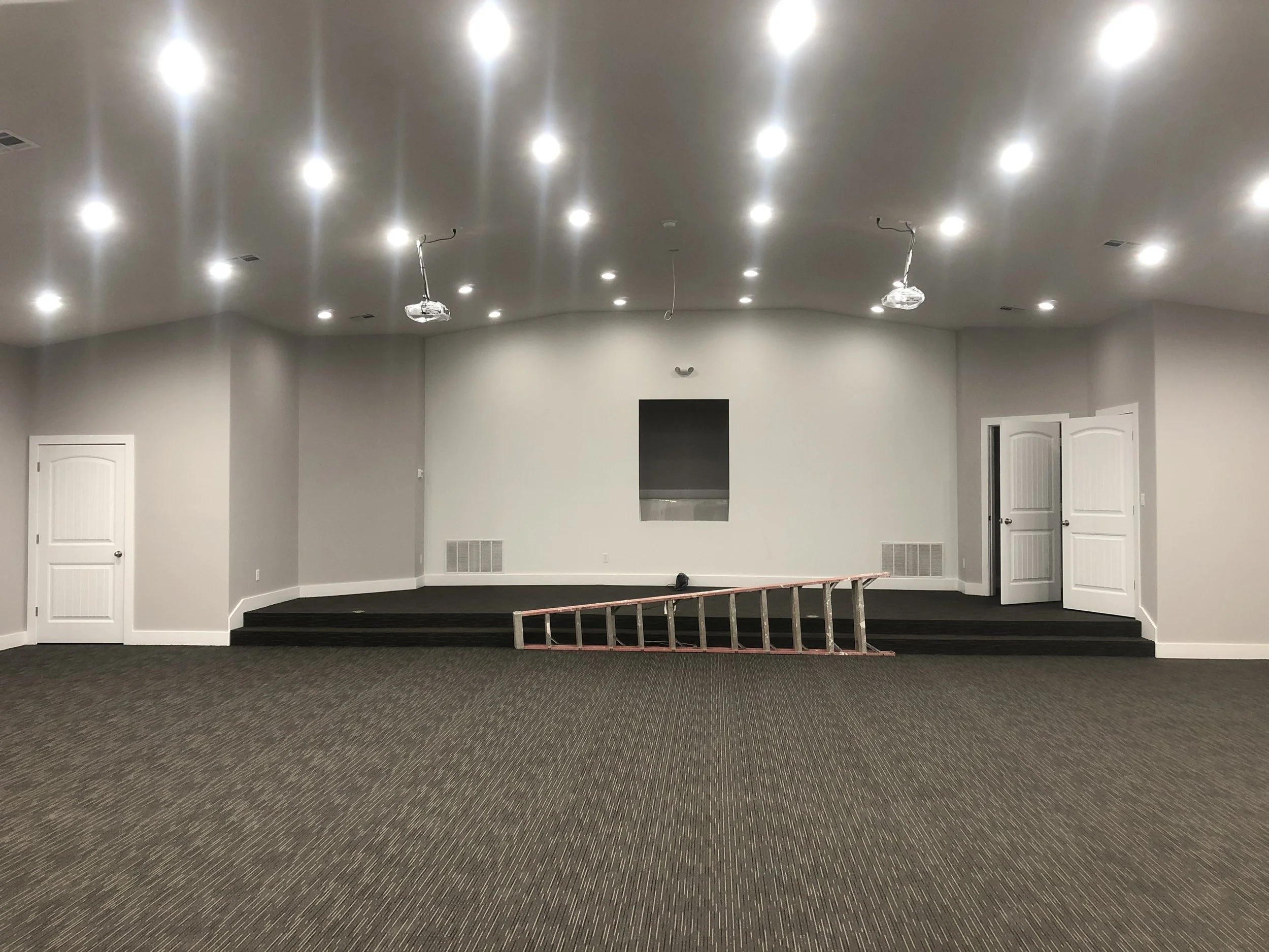 Empty room with white walls, a stage with black carpet, and multiple ceiling lights. A ladder is lying on the floor in front of the stage, and there are two doors on either side of the room.