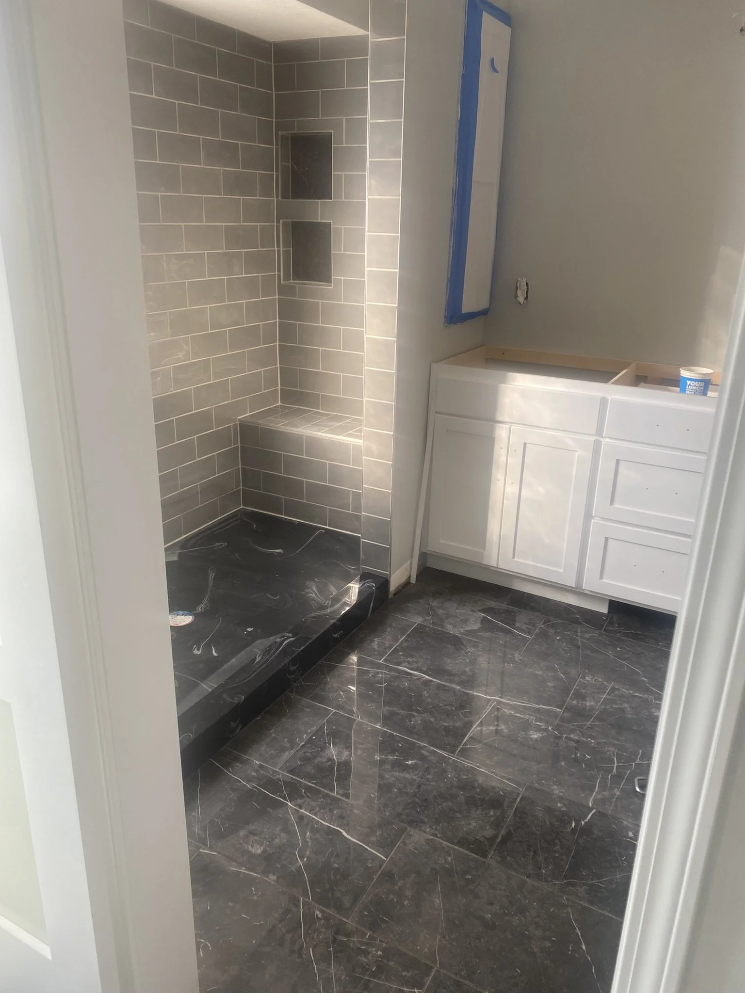Bathroom with a tiled shower area and a white vanity cabinet under construction. The shower has gray tiles and built-in shelves. The floor is tiled with dark marble-like tiles.