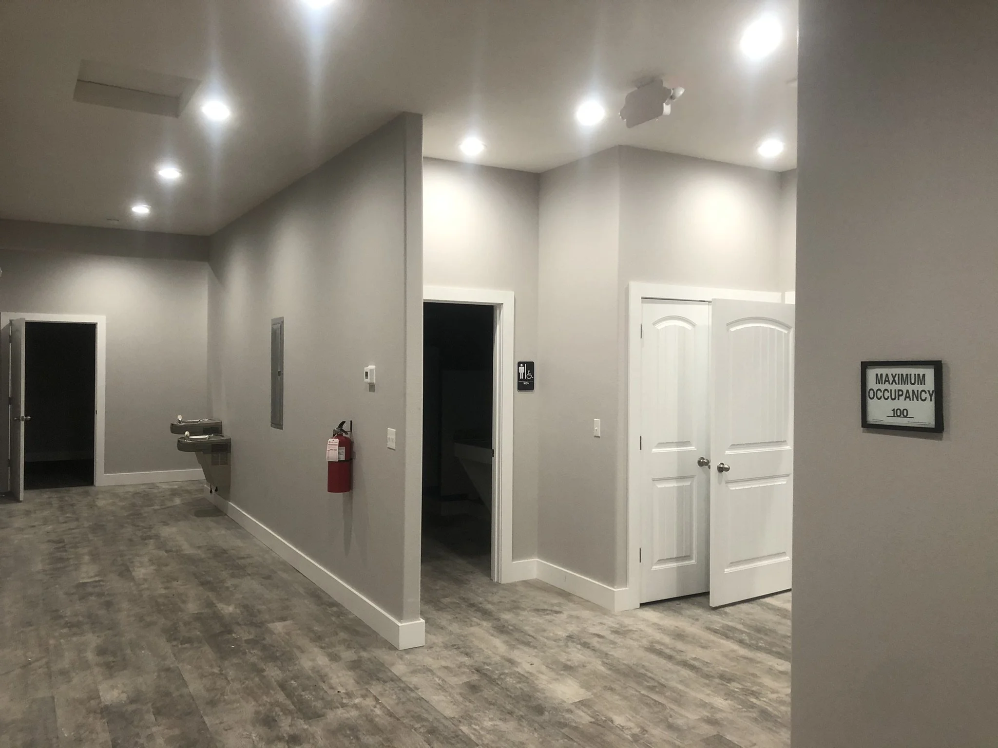 Interior view of a public restroom area with multiple doors, including men and women signage, a fire extinguisher, and a sink with a mirror, illuminated by ceiling lights.