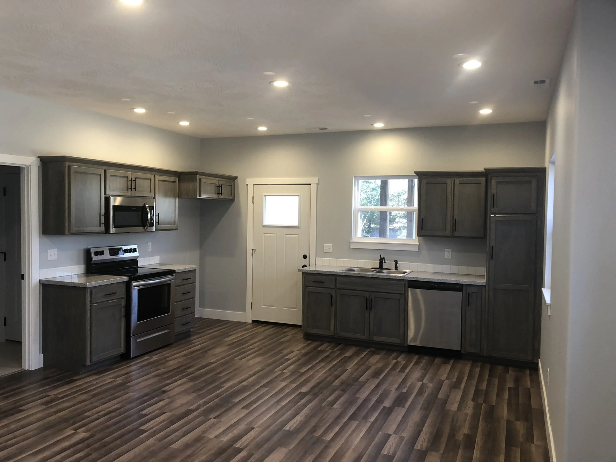 A modern kitchen with gray cabinets, a stainless steel microwave, oven, and dishwasher, a white door, a window, dark wood flooring, and recessed ceiling lights.