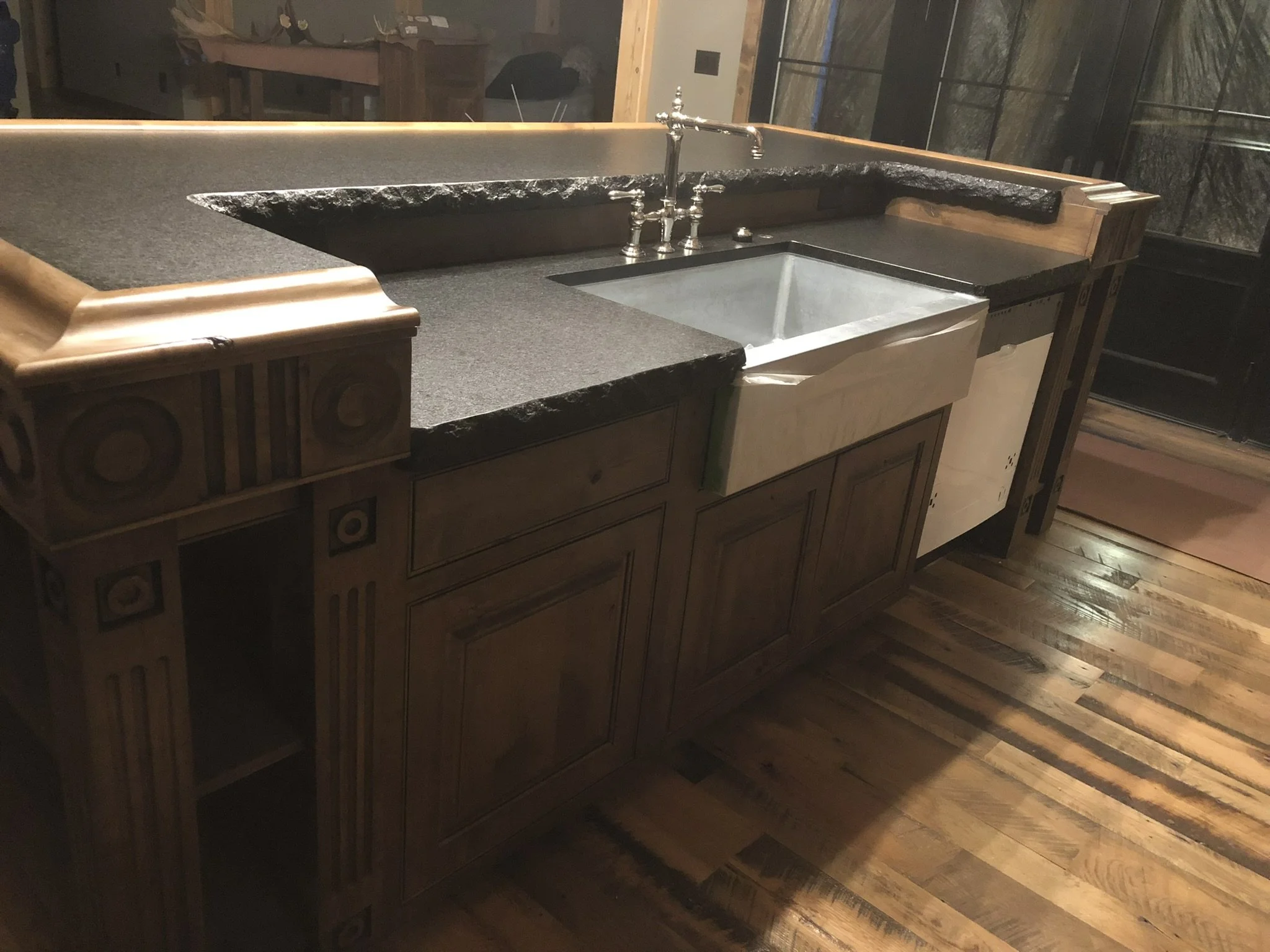 Kitchen island with a black stone countertop, a stainless steel sink, and a vintage-style faucet. Wooden cabinetry with decorative carvings and a window with a view of trees outside.