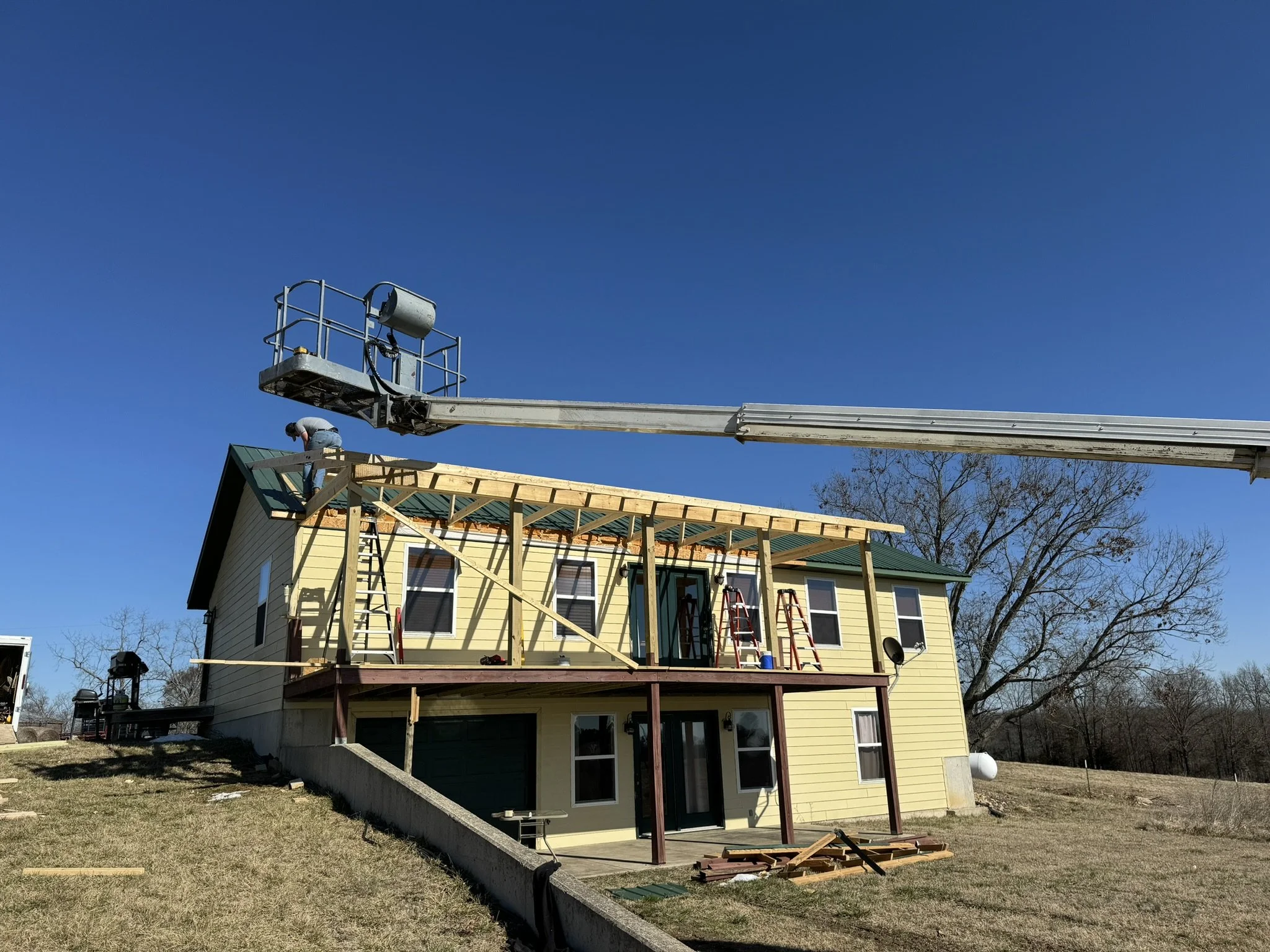 A house under construction with a yellow exterior, a green roof, and an unfinished wooden deck. A worker is on an elevated lift working on the roof's structure, and construction materials are scattered around.