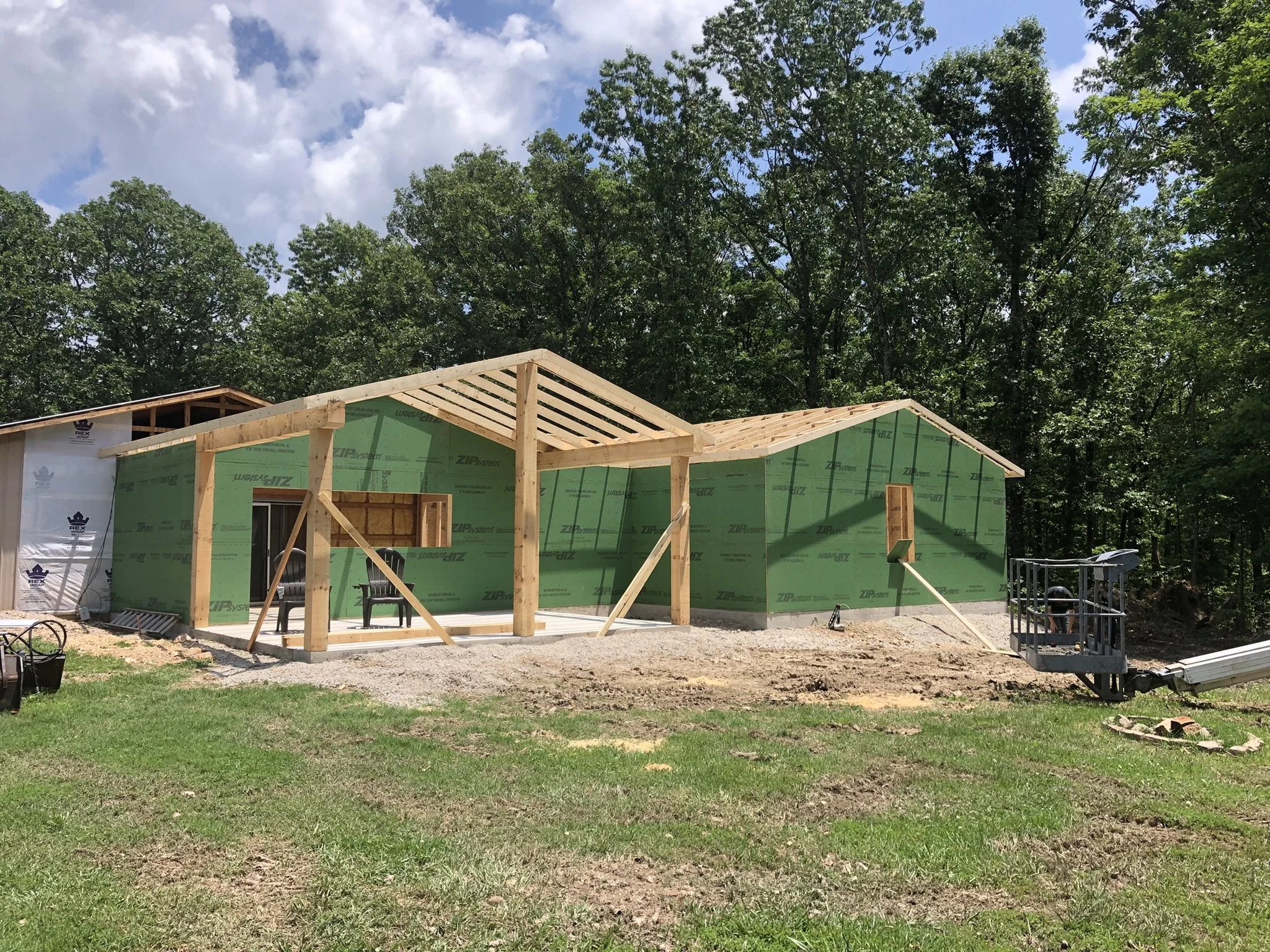 Construction site of a house in progress with partly finished wooden frame and green house wrap, surrounded by trees and cloudy sky.