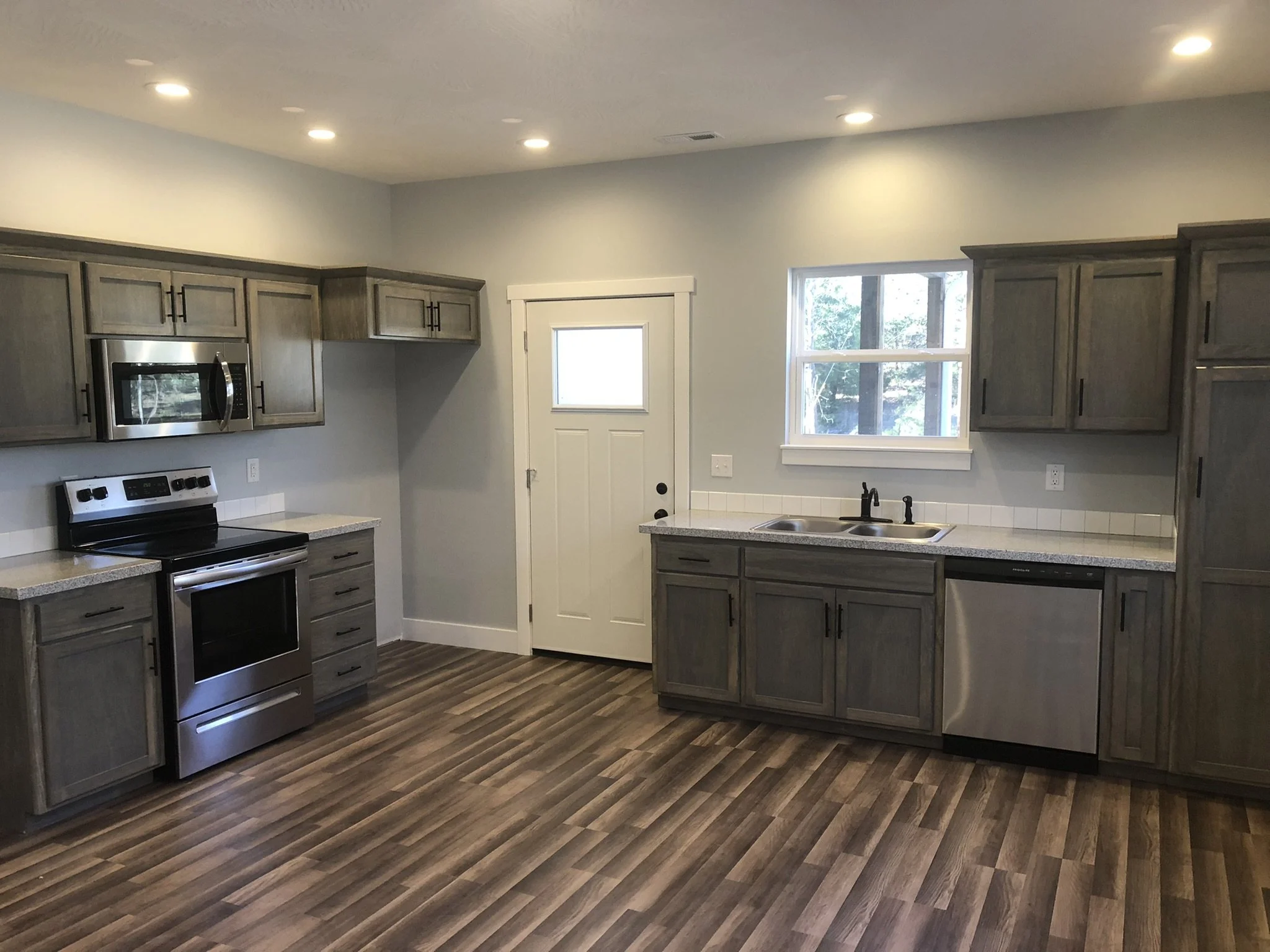 Kitchen with gray cabinets, stainless steel microwave and stove, two windows, wood-looking floor, and white walls.
